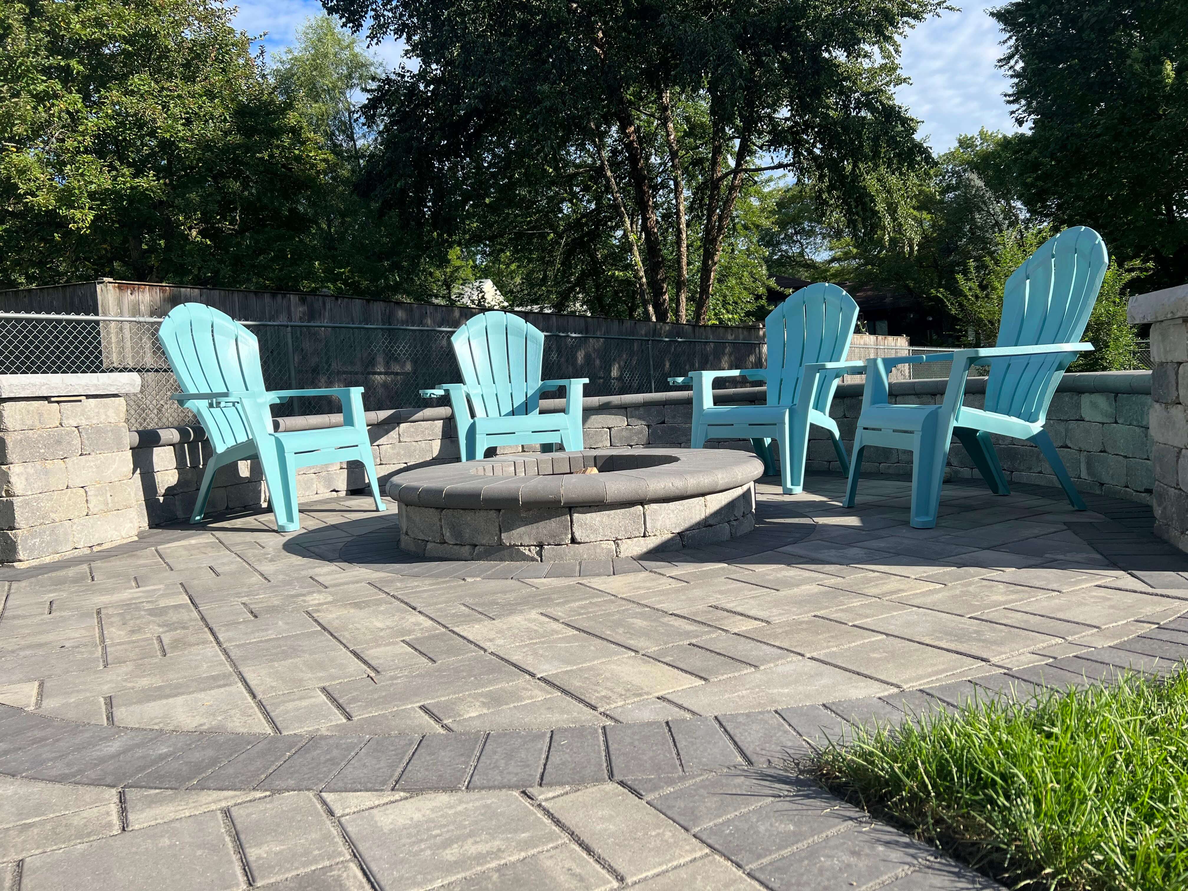 Circular stone firepit surrounded by blue Adirondack chairs on gray brick patio with a stone retaining wall.