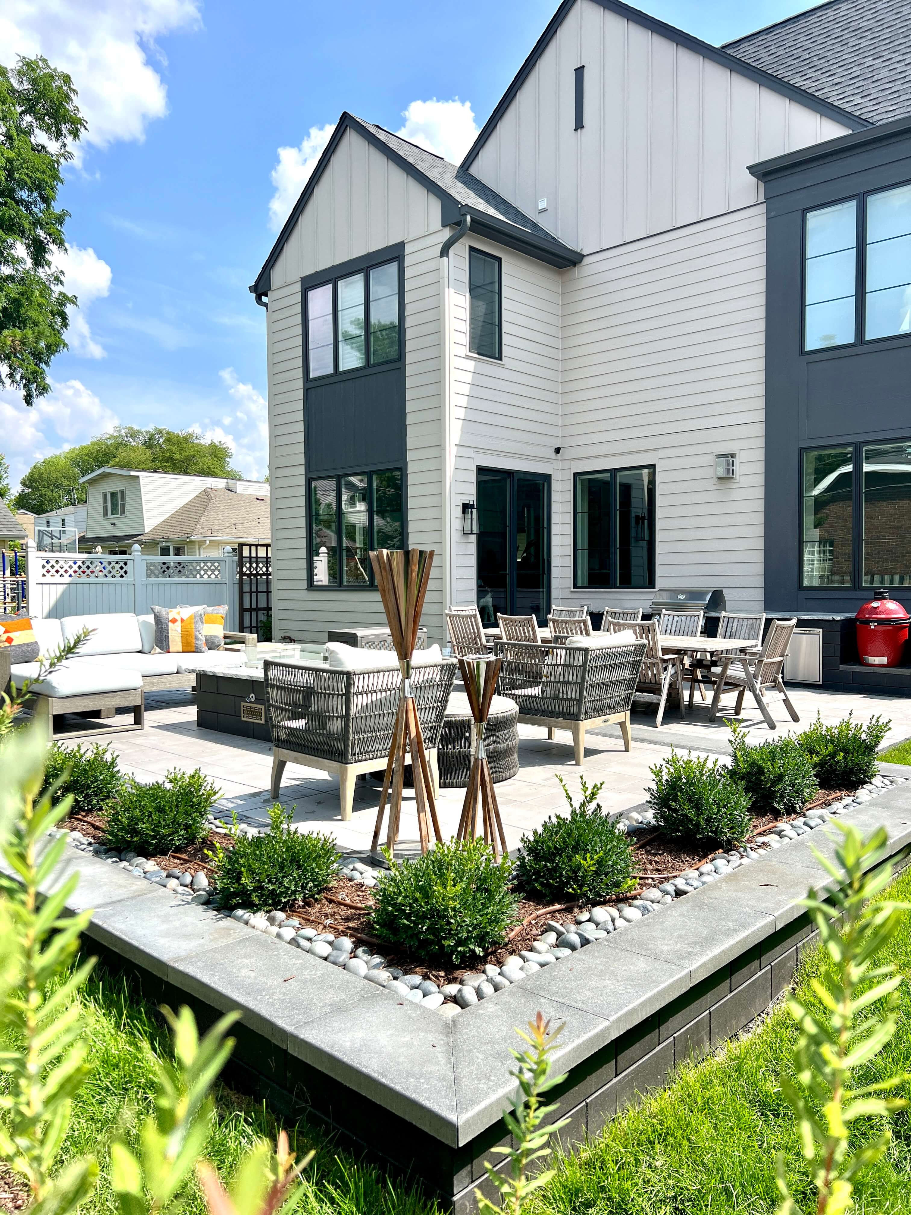 A modern house backyard with two patio areas including loungers, dining table, outdoor kitchen, and greenery.