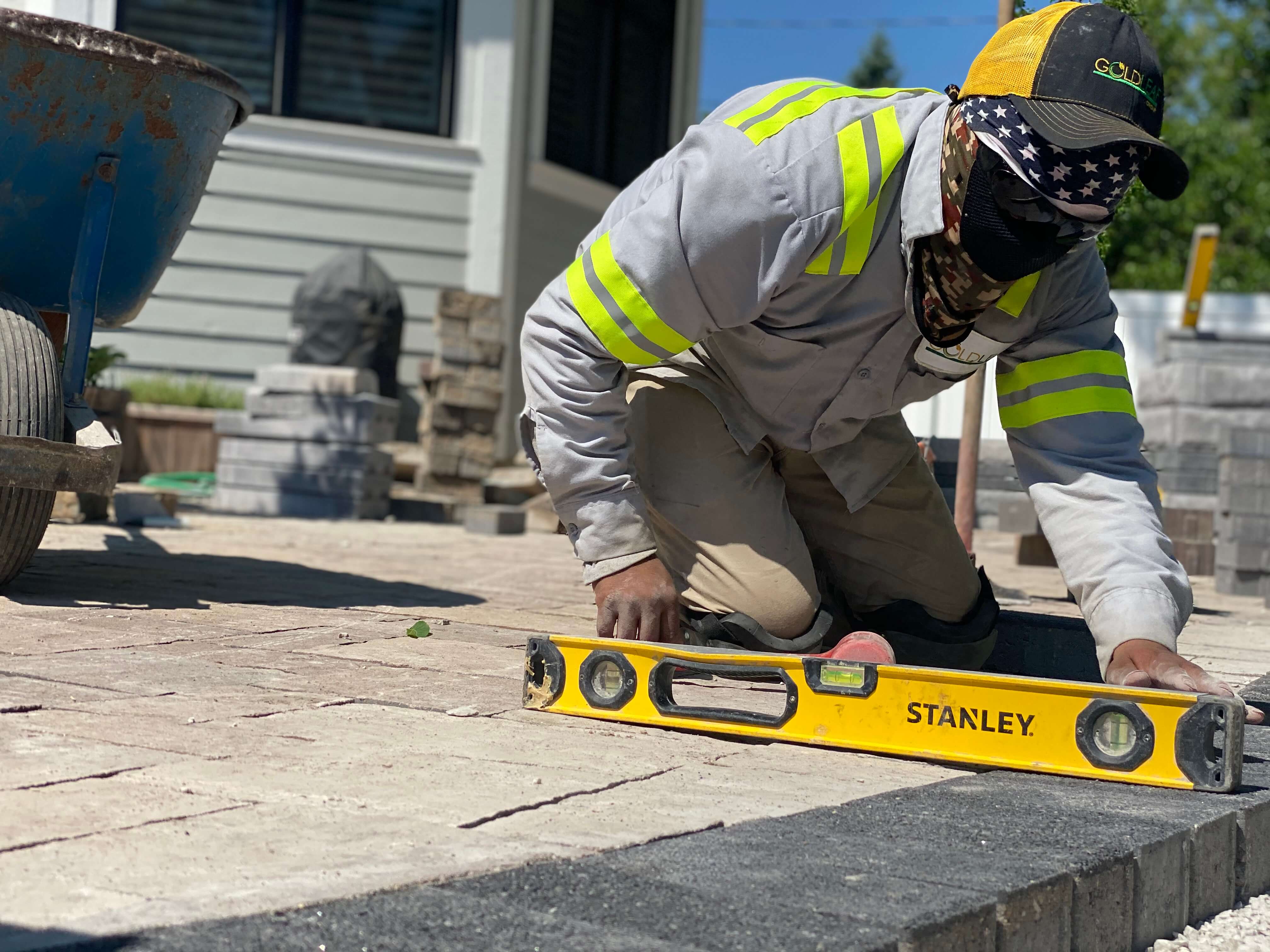 Construction worker in safety gear using a yellow level tool to align stone pavers outdoors.