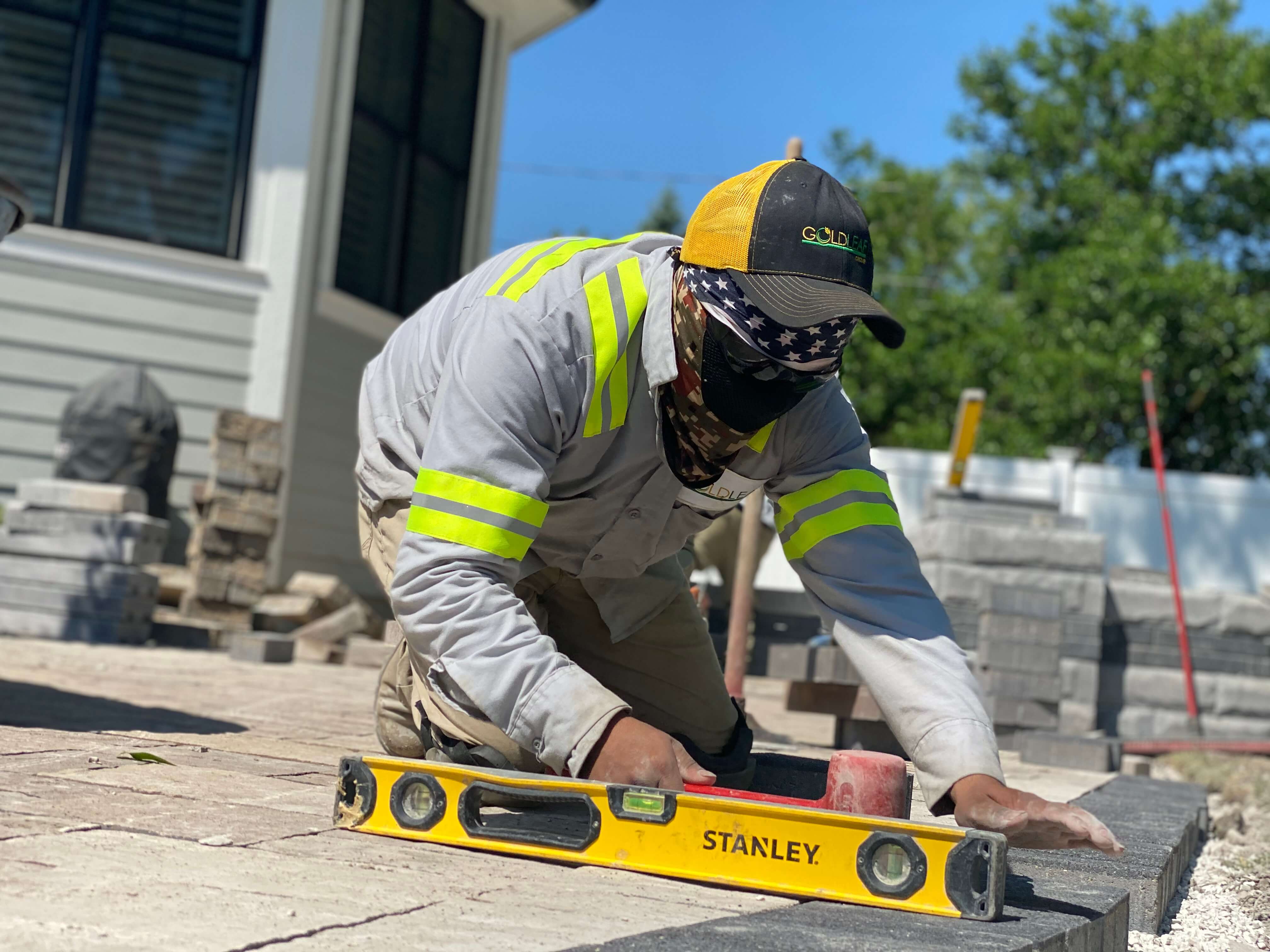 Worker wearing a mask and cap using a yellow level tool to lay bricks on a sunny day.