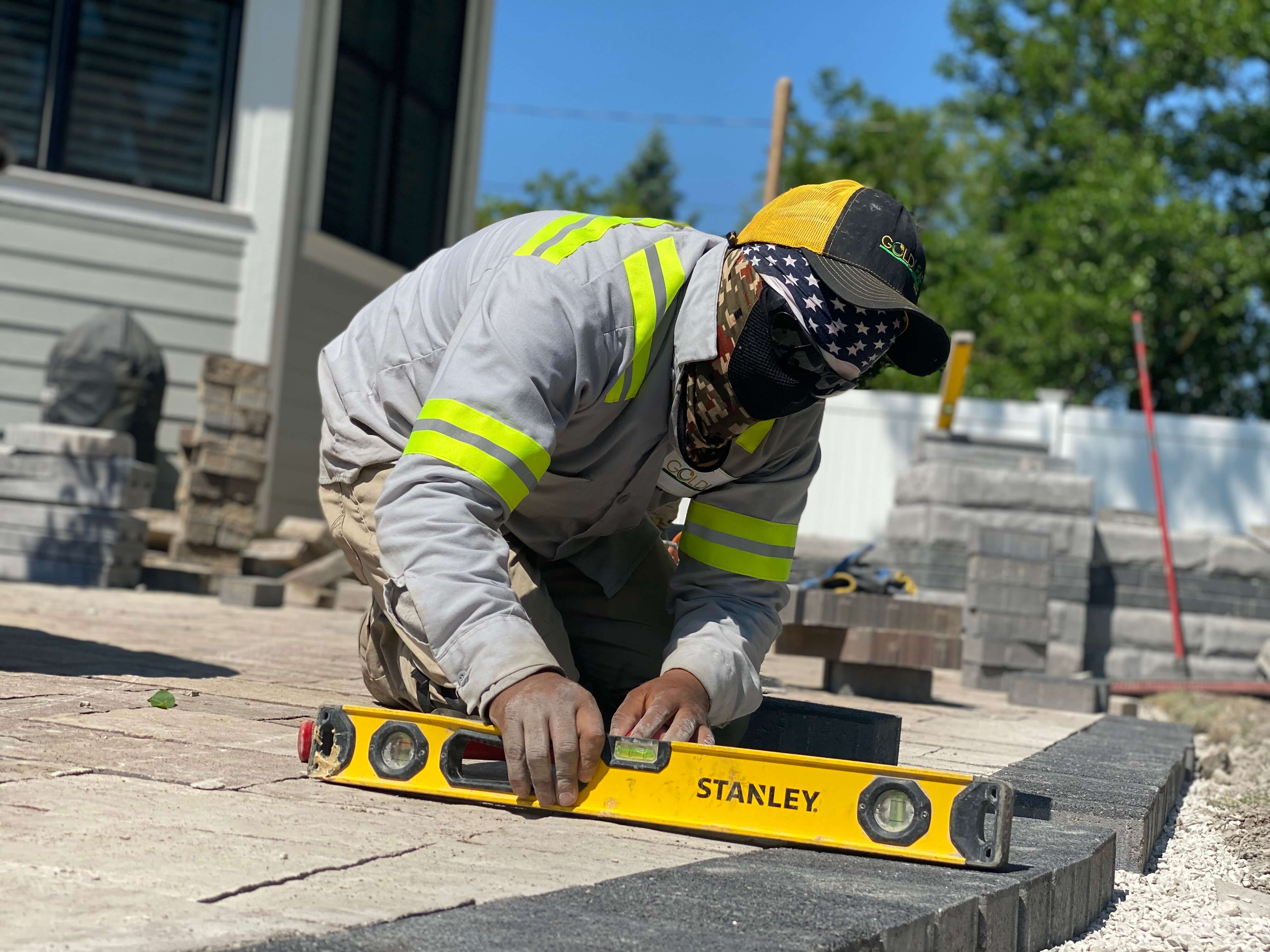 Worker wearing reflective shirt and cap uses a level tool to install brick pavers outdoors.