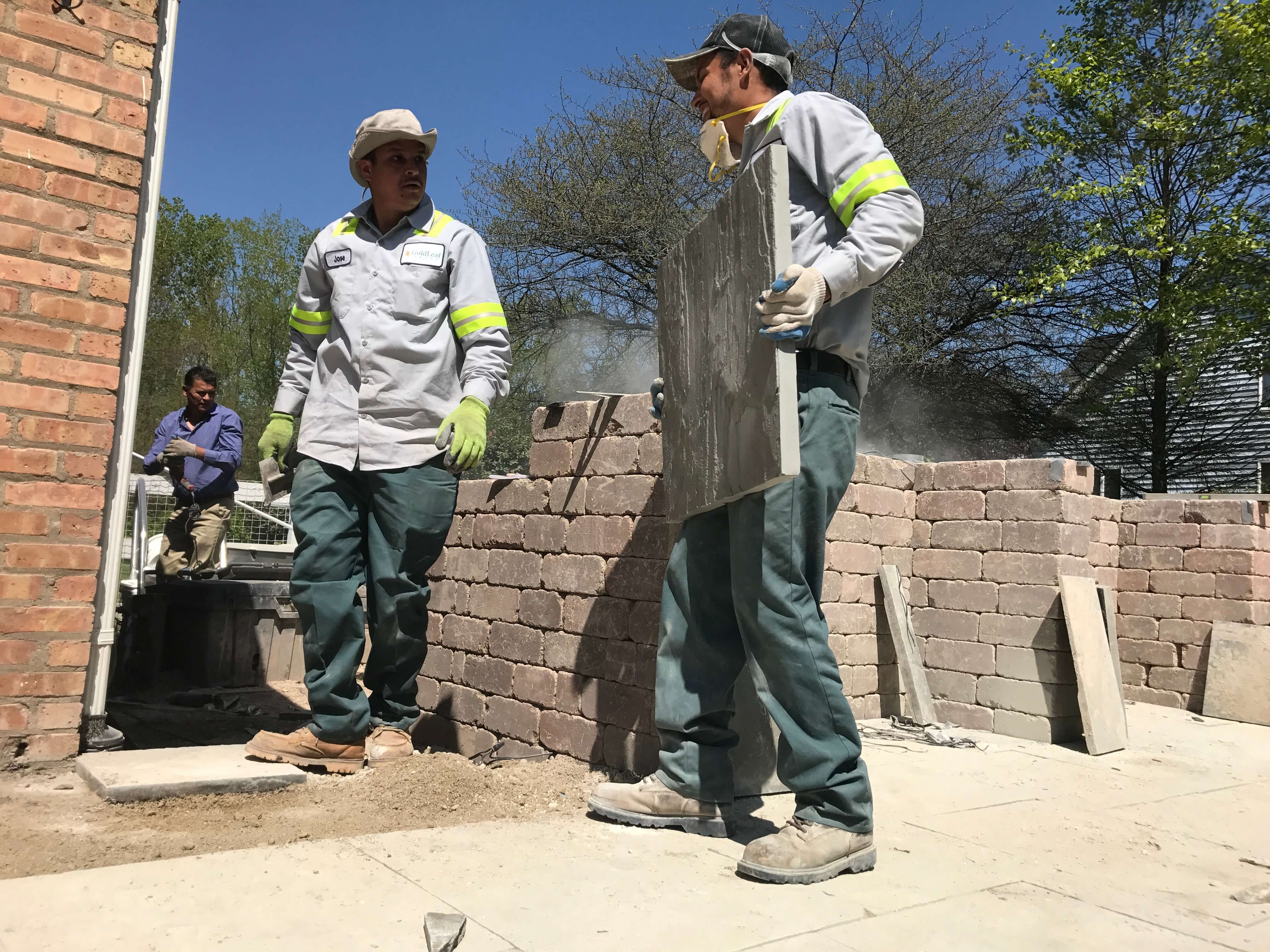 Two construction workers in uniforms building a stone retaining wall outdoors under a clear blue sky.