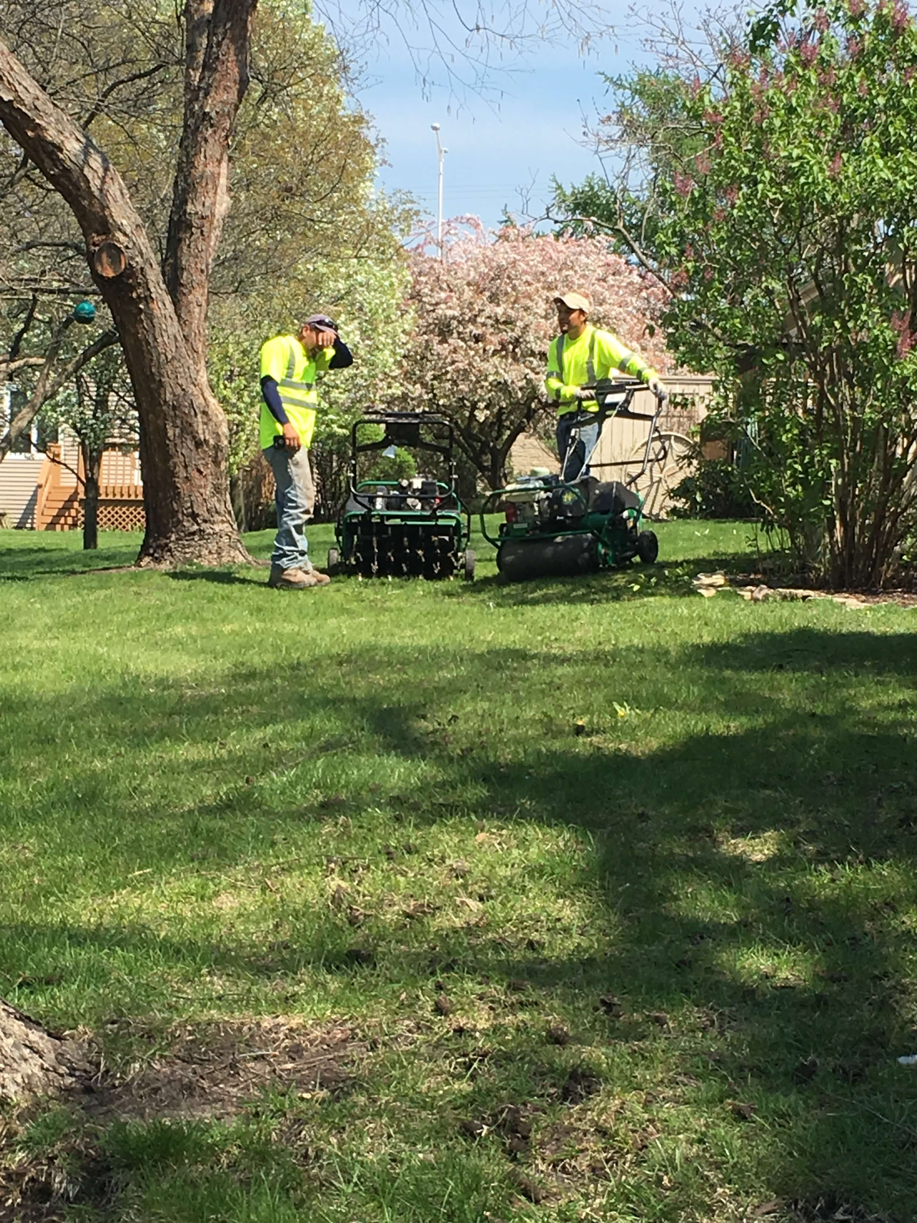 Two workers in yellow shirts operating lawn aeration machines on a green grassy yard.
