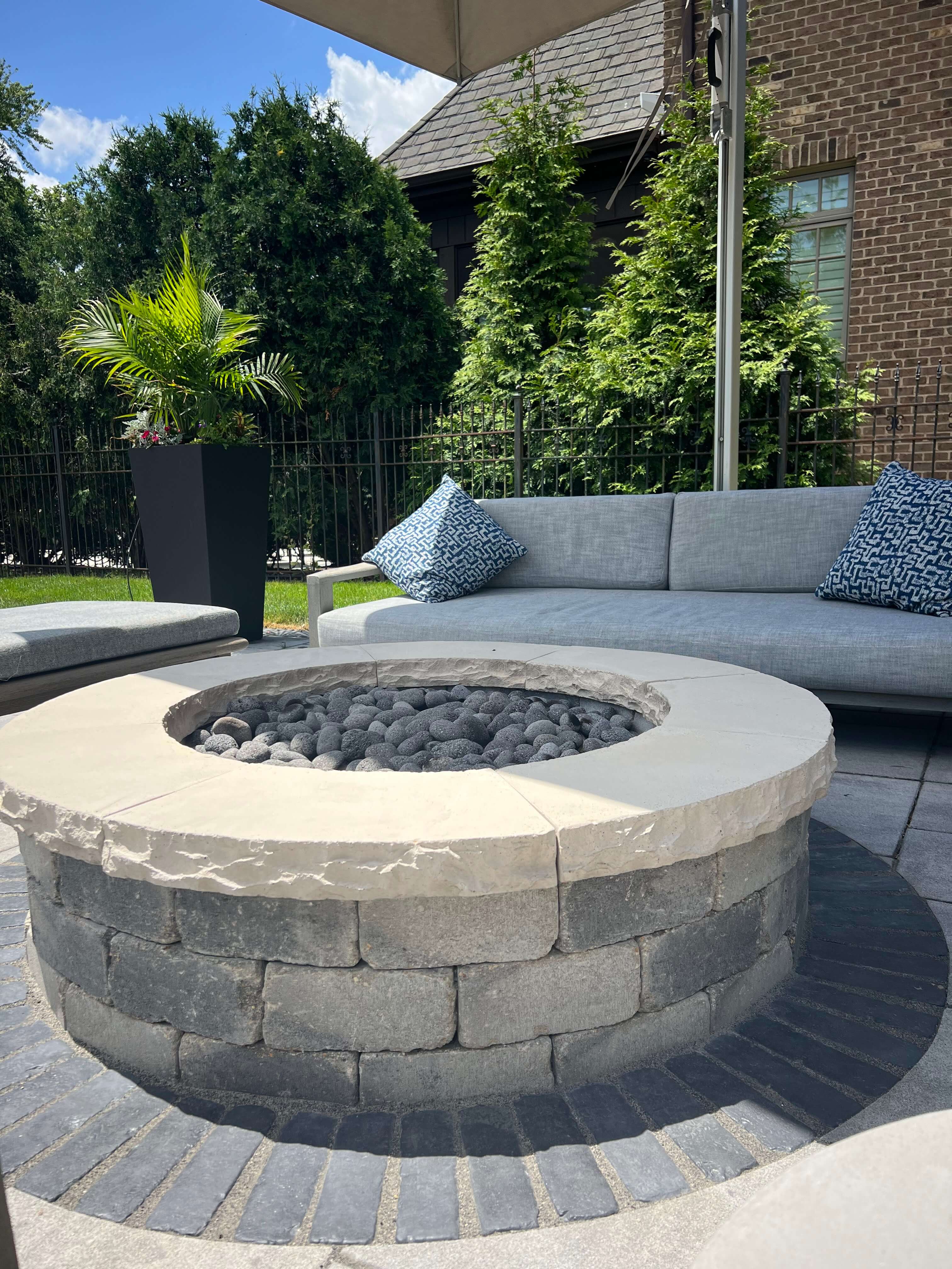 Outdoor patio with gray cushioned chairs around a stone fire pit beneath a large umbrella.