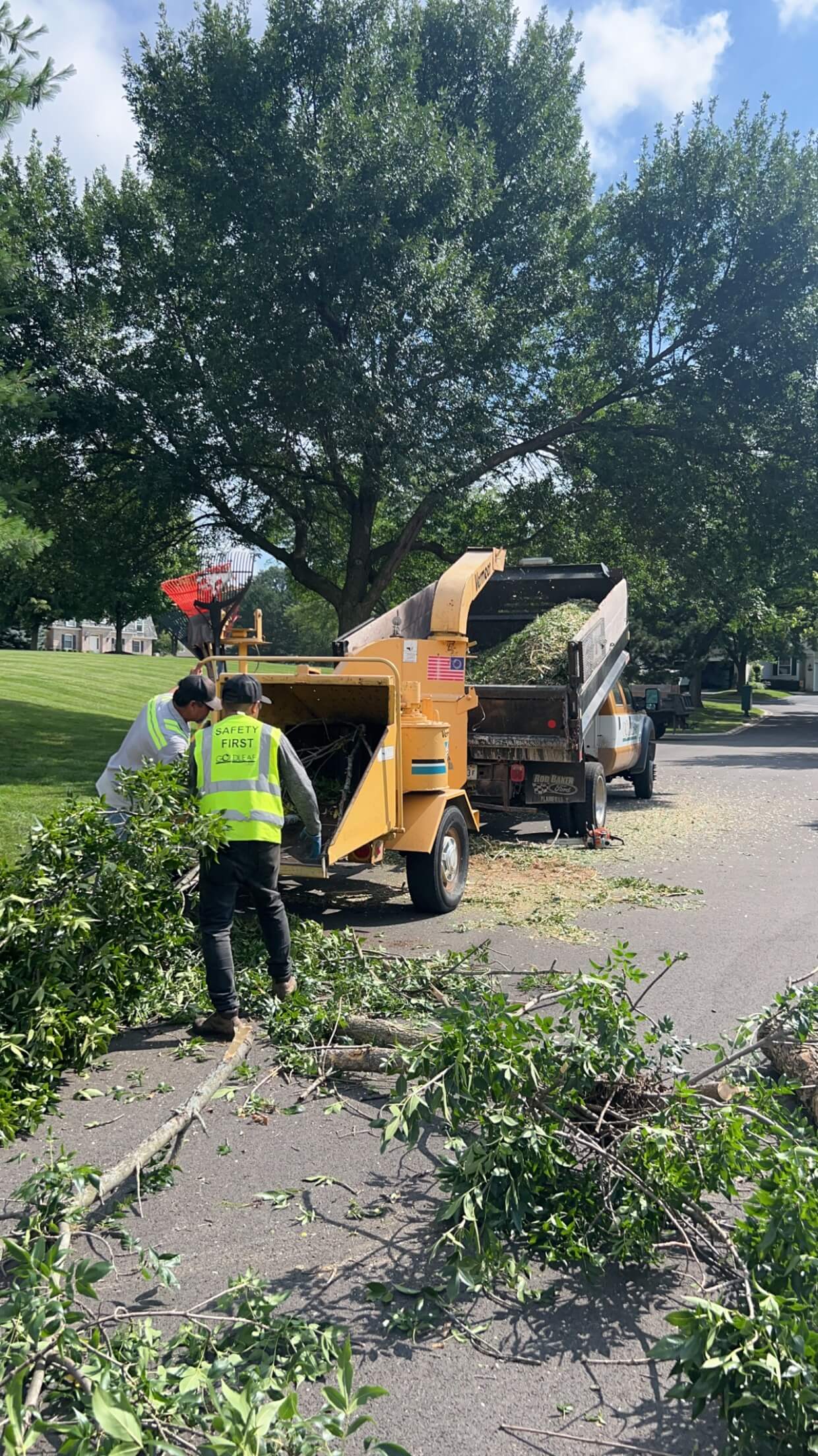 Two workers feed tree branches into a yellow chipper with a truck collecting green debris on a street.