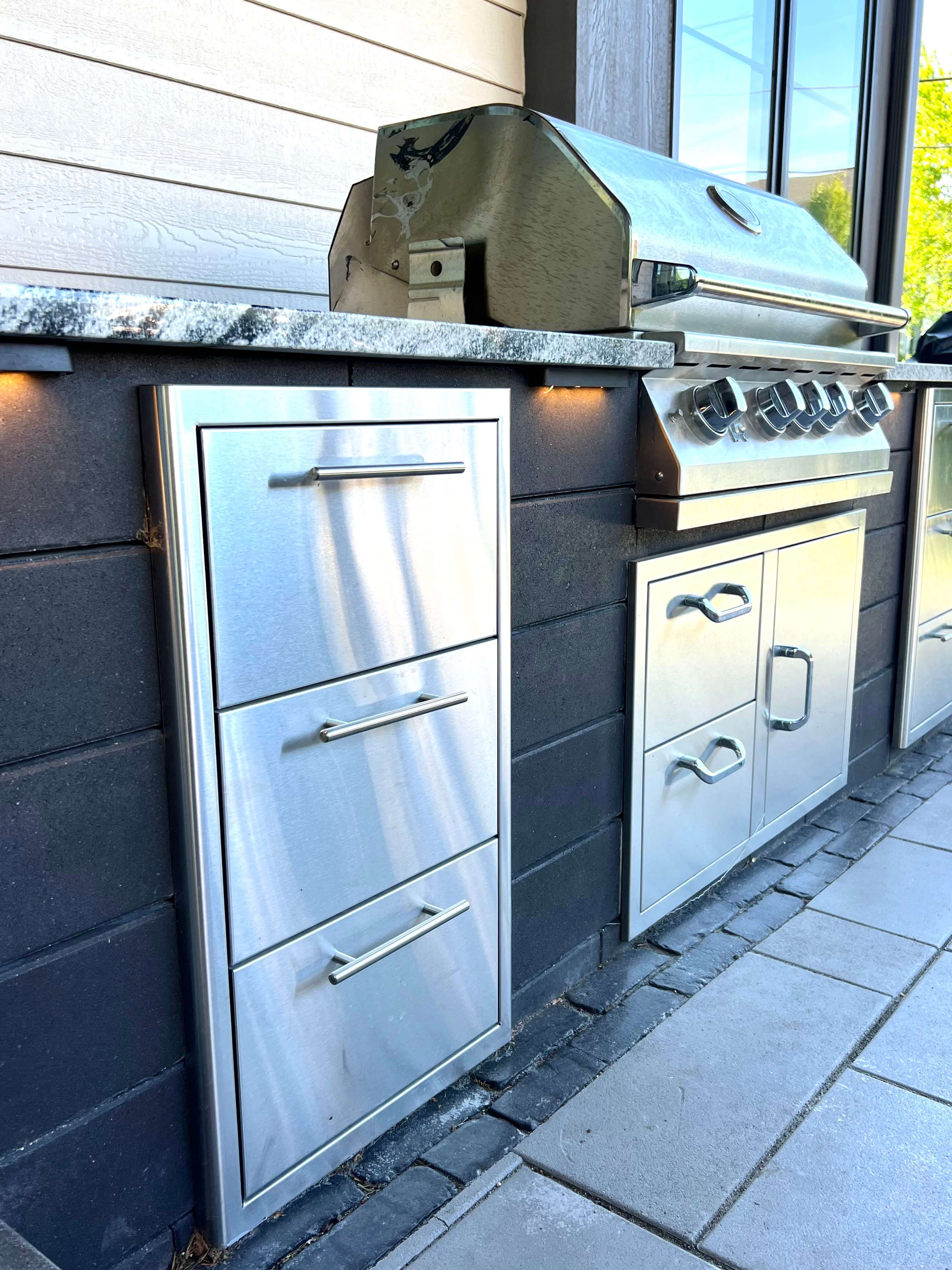 Outdoor kitchen with stainless steel grill and storage drawers set in a stone counter and dark brick base.