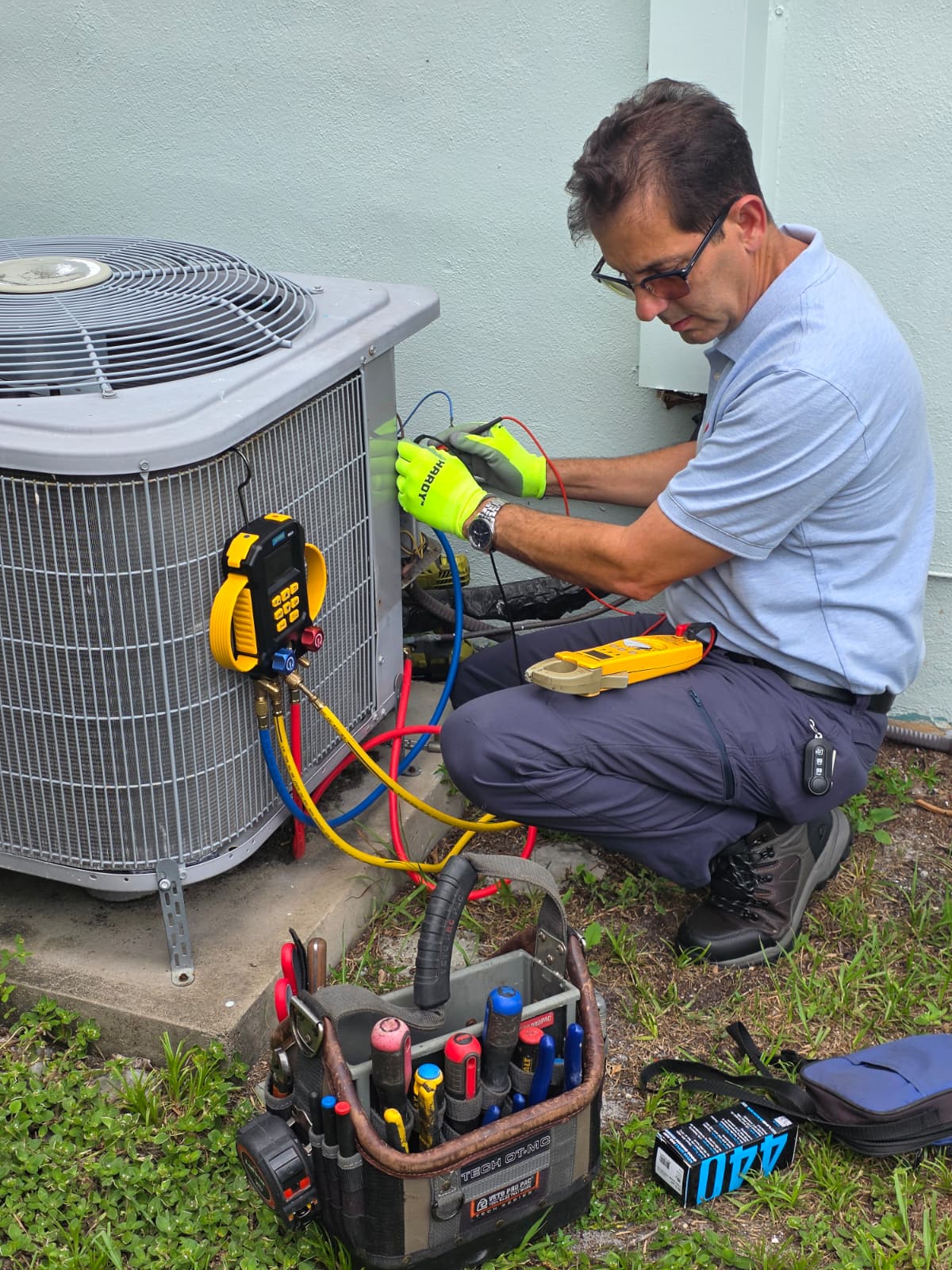 Technician wearing gloves fixing an outdoor air conditioning unit with tools and gauges nearby.