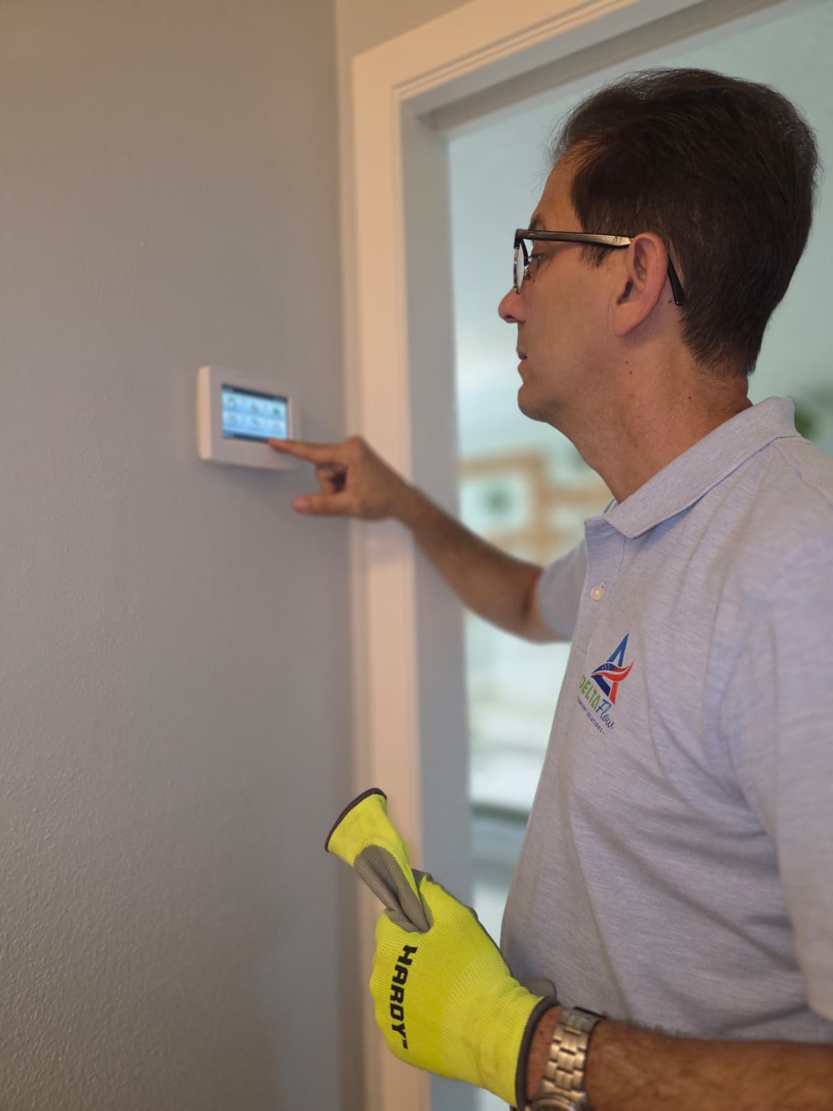 Man wearing glasses and a glove adjusting a wall-mounted touchscreen thermostat in a home.