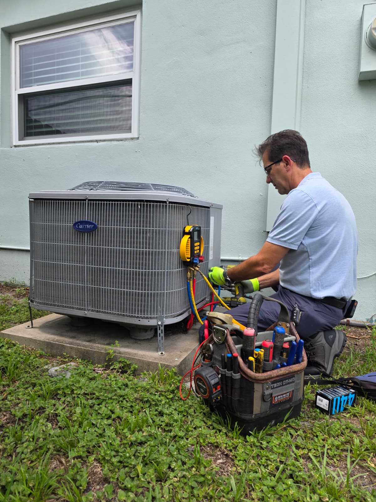 Technician in gloves repairs an outdoor Carrier air conditioning unit with tools on grass.