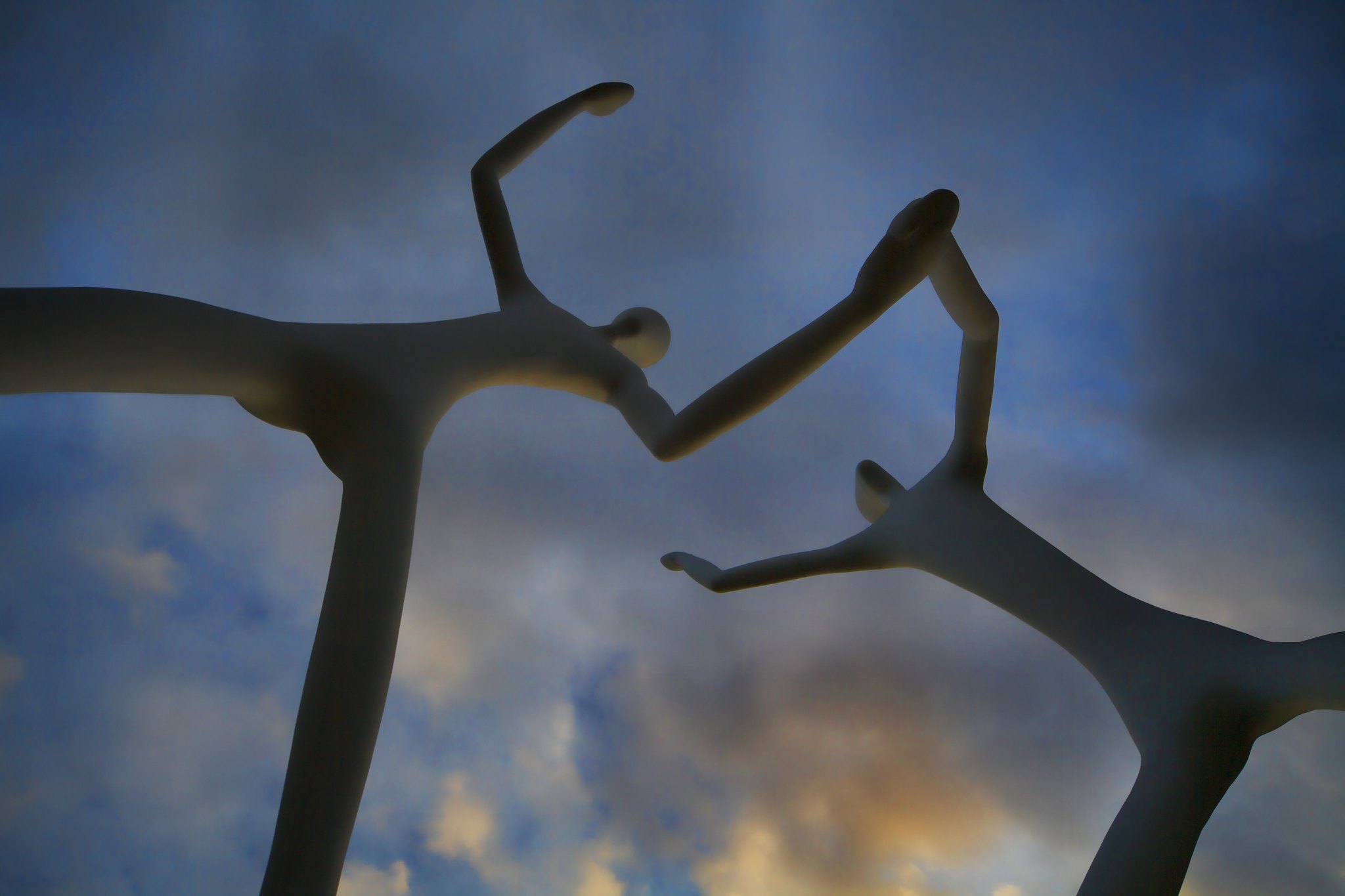 A photo of tall slim humanoid statues holding hands and dancing against a cloudy sky.