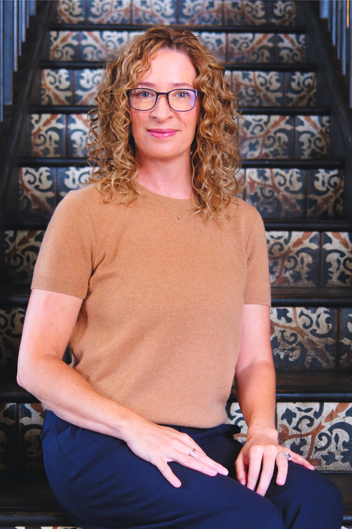 A photo portrait of Erin Bunyard, a white woman with dark blond curly hair, wearing a camel sweater and navy pants, sitting on steps made of decorative tile.