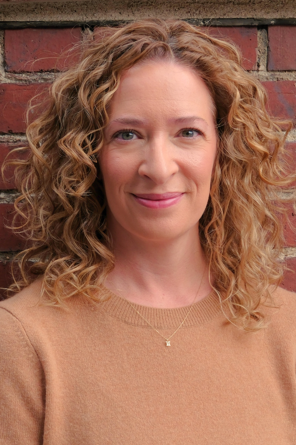 A photo portrait of Erin Bunyard, a white woman with dark blond curly hair, wearing a camel sweater, against a brick background.
