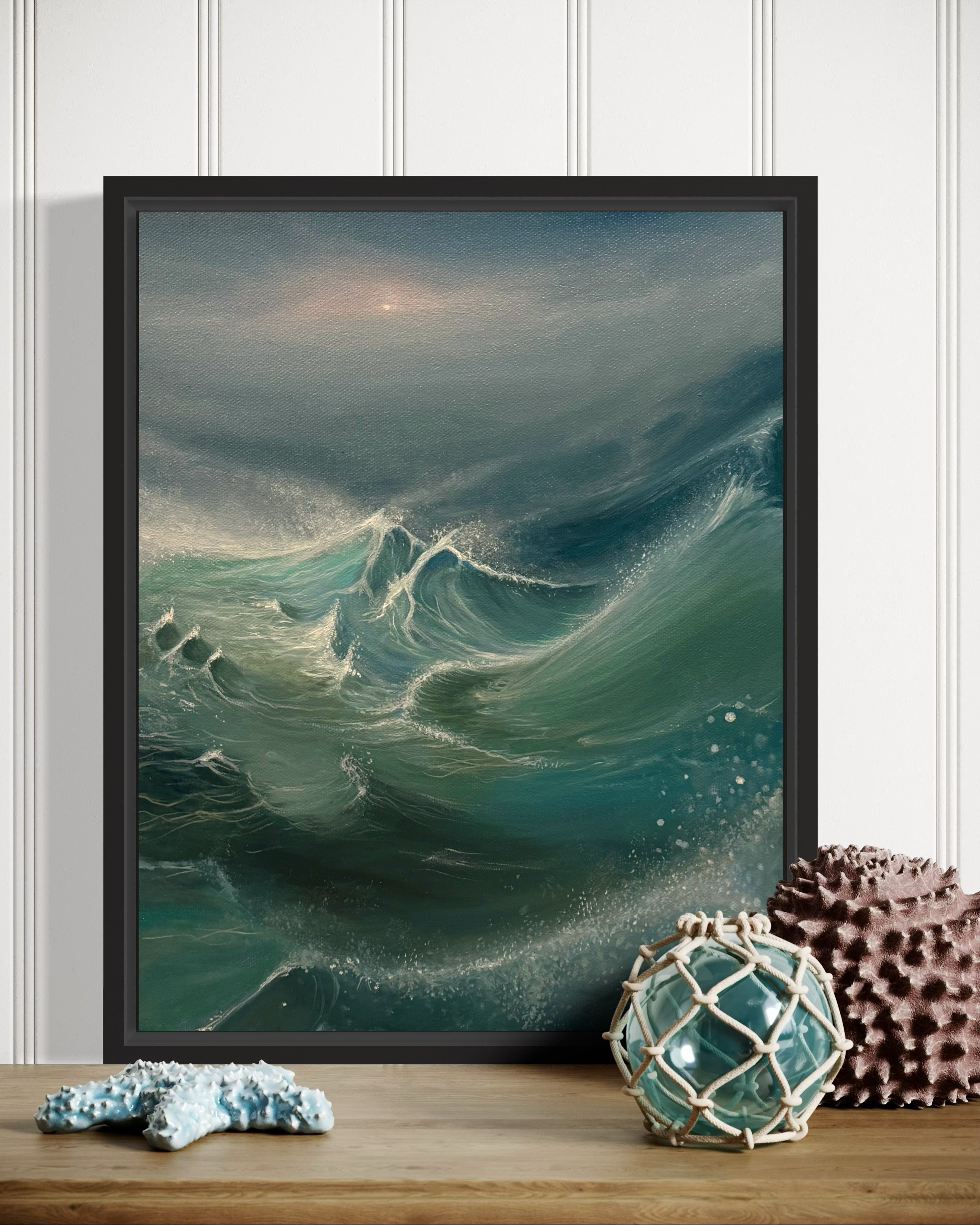 A framed print of one of Lara Moriarity's seascape paintings, propped on a wooden shelf beside shells and a glass float