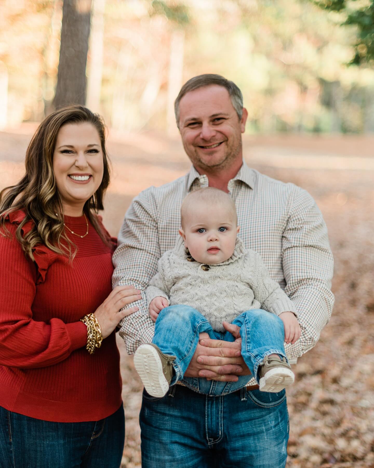 Drew and family smiling outdoors with the man holding a baby dressed in a sweater and jeans.