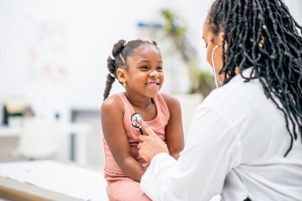 Female doctor using a stethoscope to examine a smiling young Black girl during a medical checkup.
