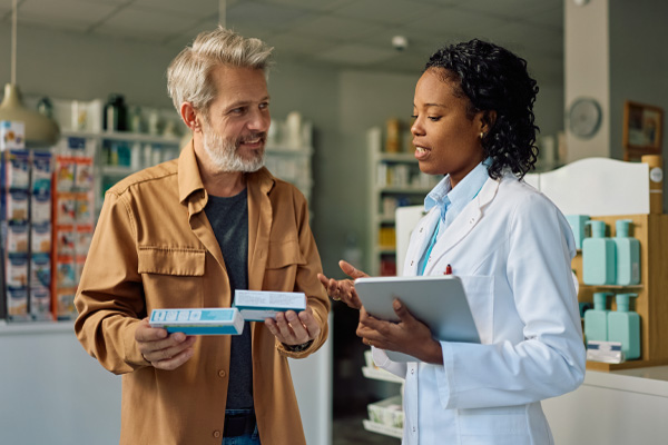 Female Black pharmacist with a digital tablet consulting a male patient about his medication in a pharmacy setting.