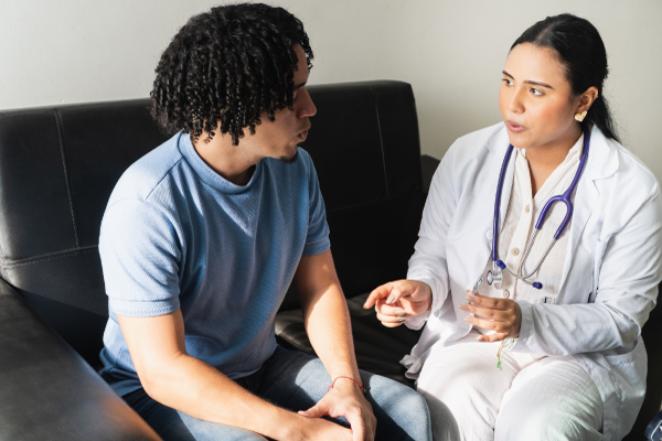 Female doctor wearing a white coat and stethoscope speaking attentively with a young male patient.