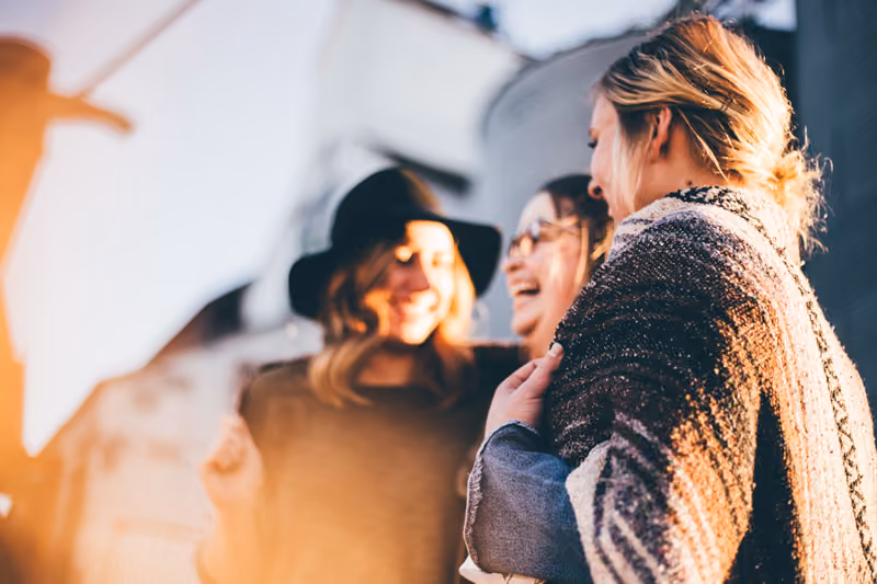Three women outdoors laughing and enjoying each other's company in soft sunlight.