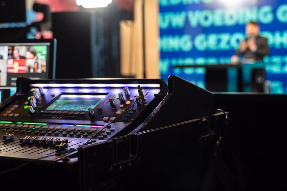 Close-up of an audio mixing console in a studio with a blurred presenter and screen in the background.