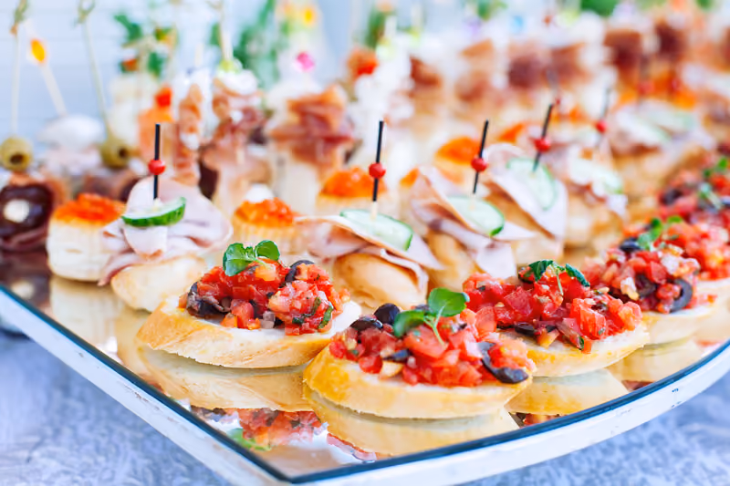 Assorted finger sandwiches and bruschetta with tomato and herbs arranged on a mirrored tray.