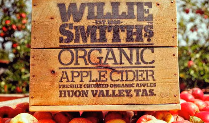 Wooden crate labeled Willie Smith's Organic Apple Cider on apples with blurred orchard background.