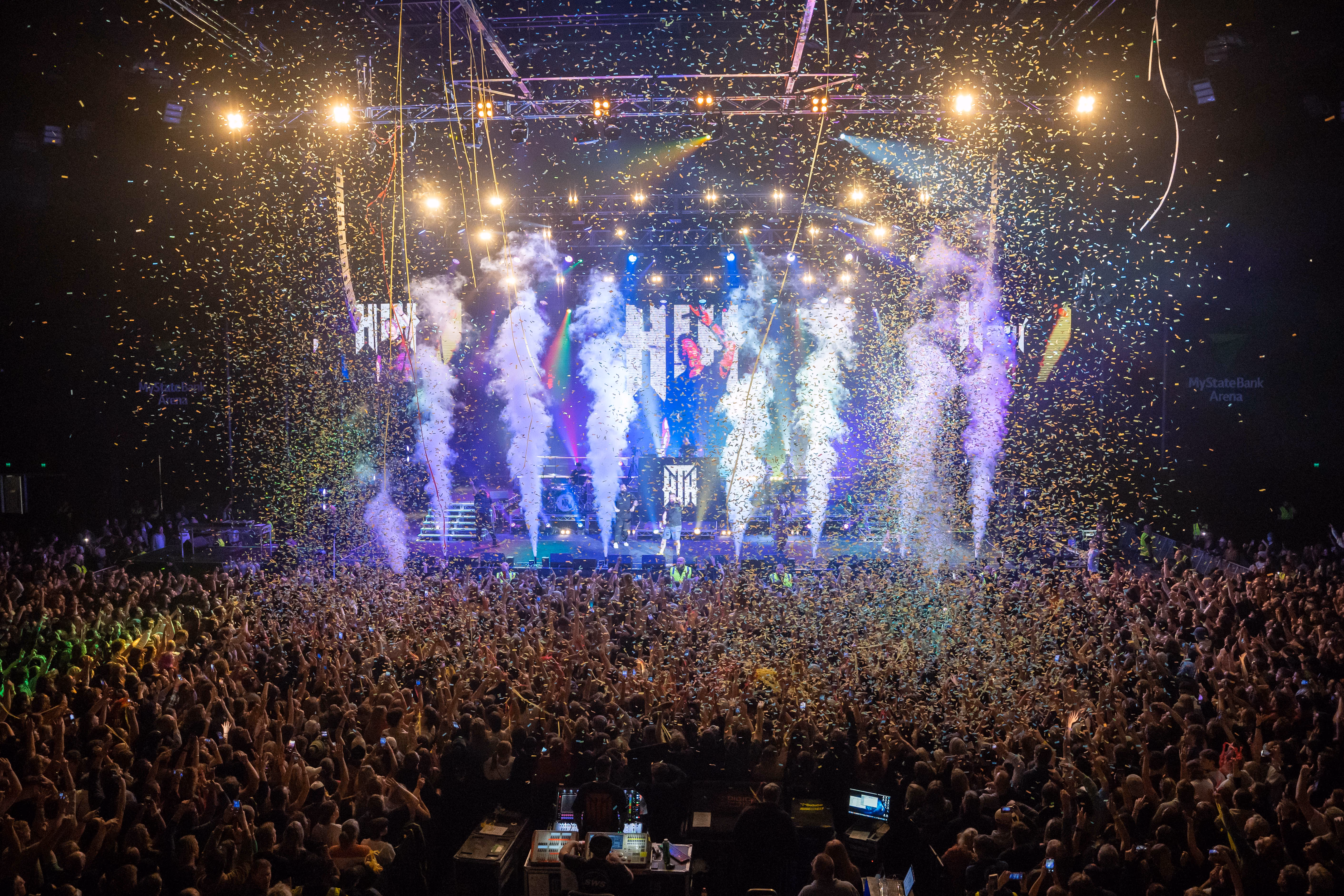 Crowded concert with stage lights, smoke effects, confetti, and a large enthusiastic audience.