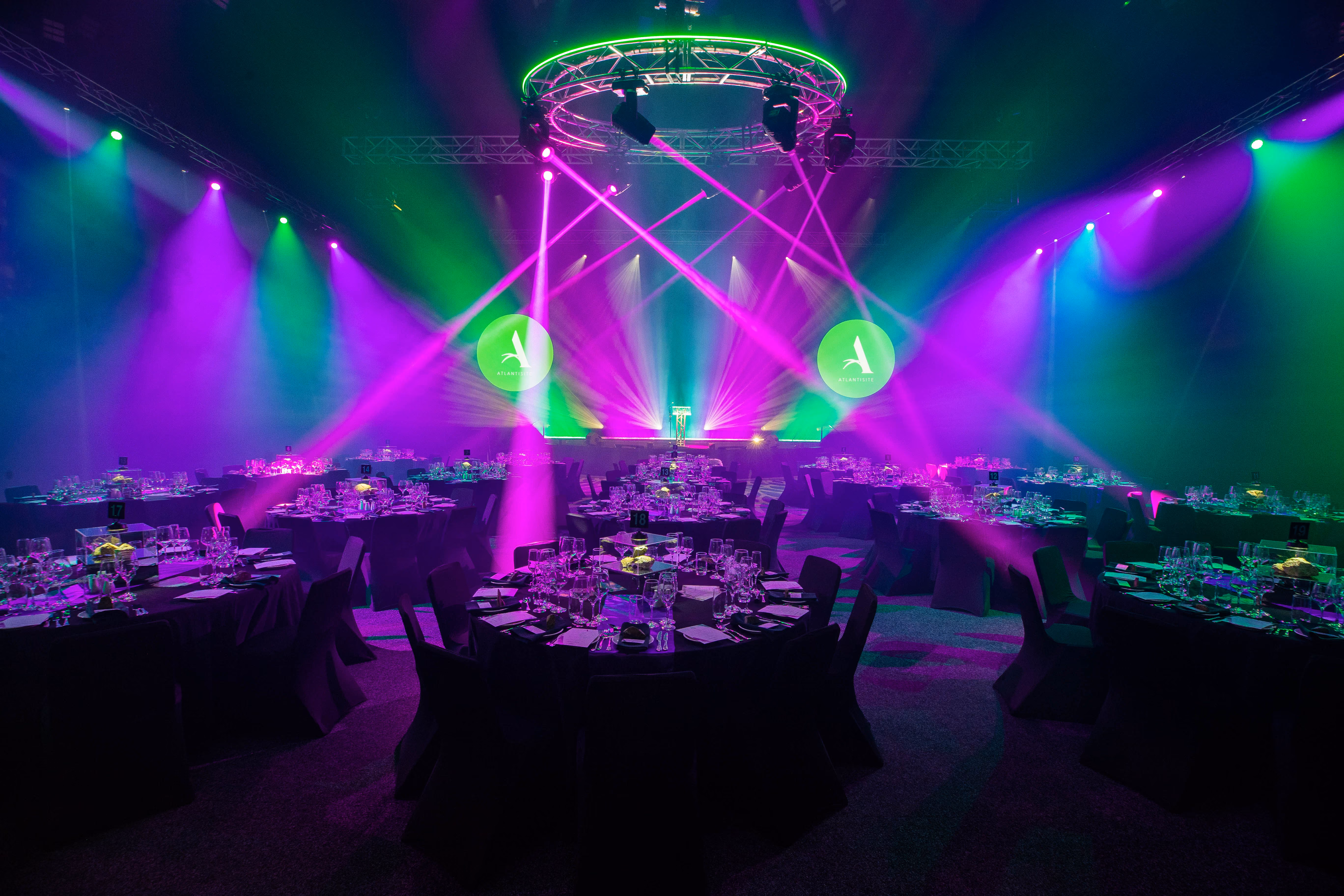 Event hall with round tables set for dinner under green and purple stage lighting.