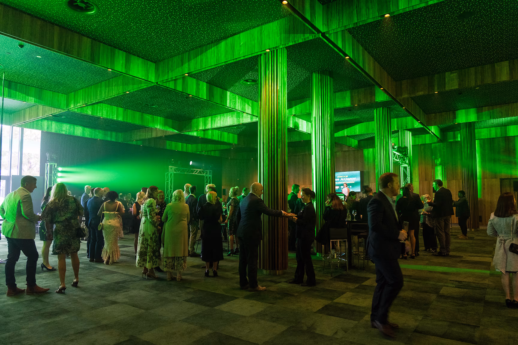 People mingling in a green-lit room with wooden pillars and a large window on the left.