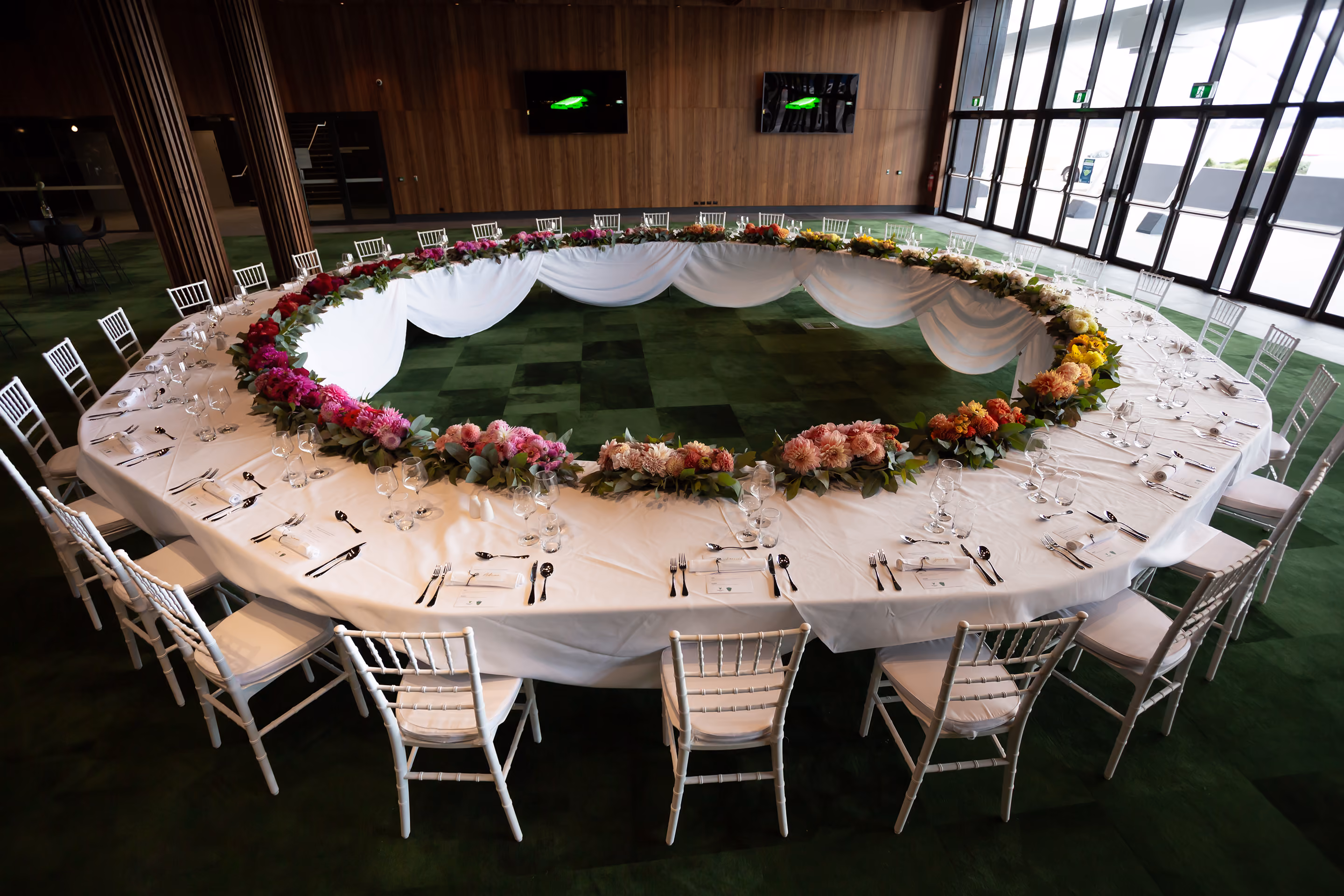 Large circular banquet table with white chairs, white cloth, and a decorative flower ring centerpiece.