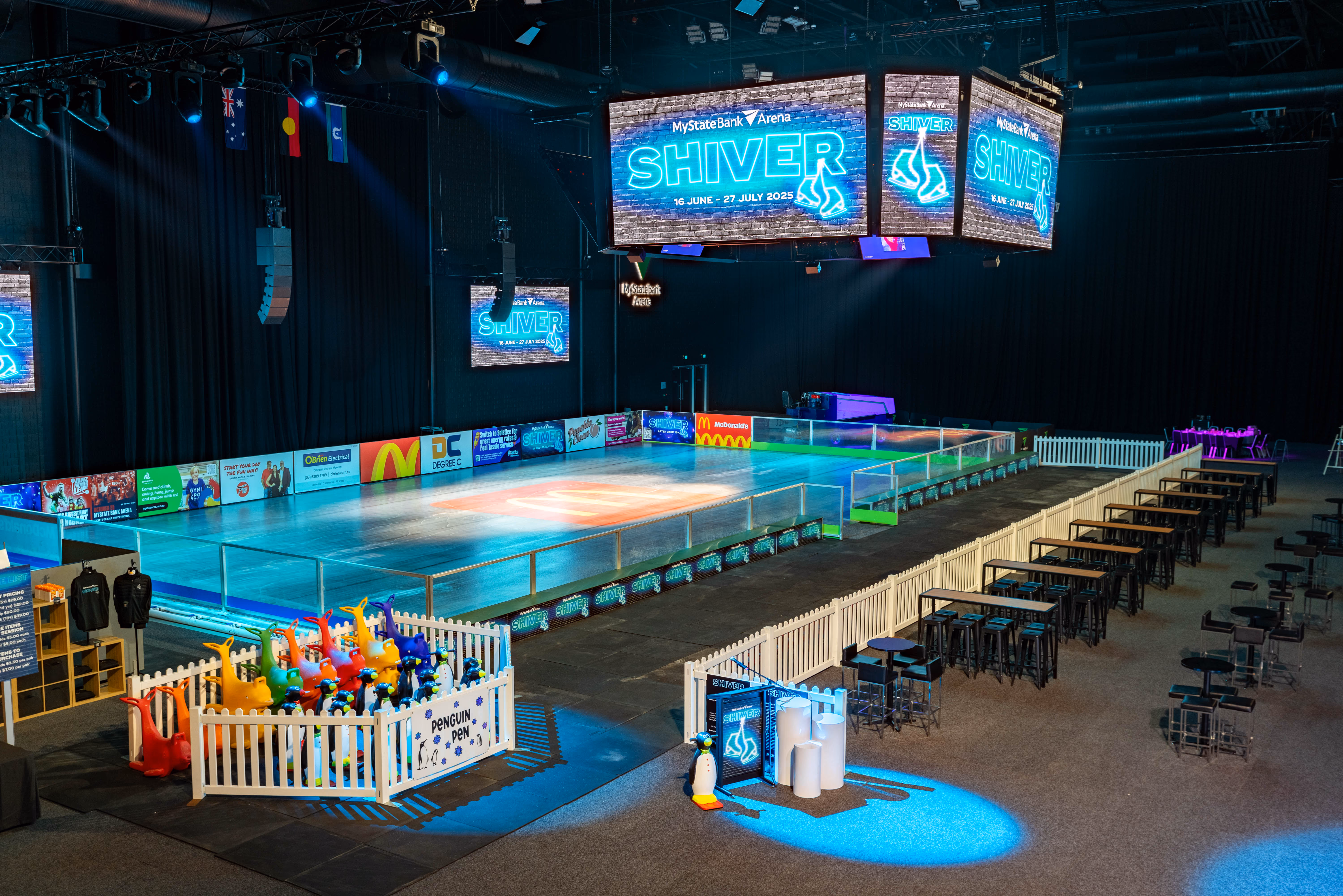 Indoor ice rink with colorful penguin skating aids, tables, and screens showing SHIVER event details.