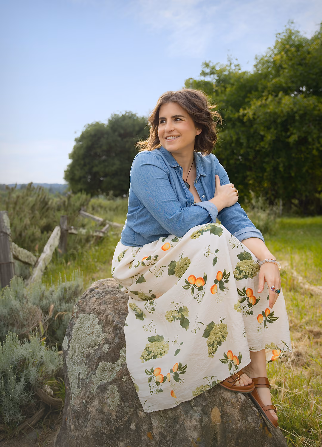 Leah Hoffman in a floral skirt and denim shirt sitting on a rock outdoors, smiling and looking to the side.