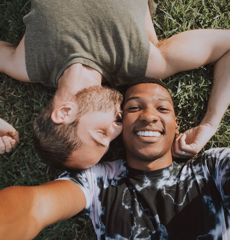 Two men lying on grass in opposite directions taking a selfie, one smiling and the other kissing his forehead.