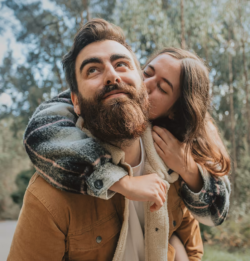 Woman hugging and kissing a smiling bearded man outdoors with trees in the background.