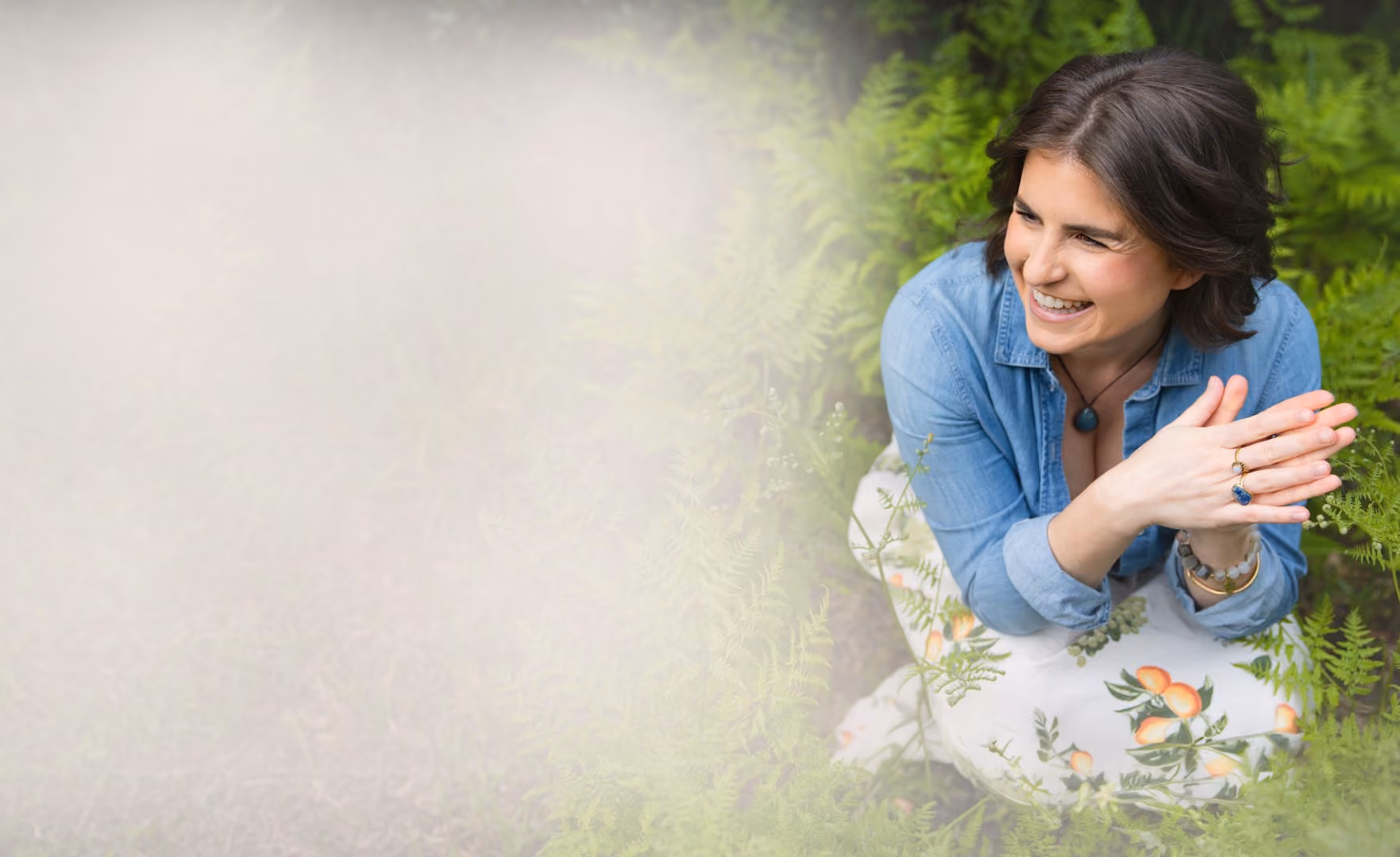 Leah Hoffman in a denim shirt and floral skirt sitting among green ferns outdoors.