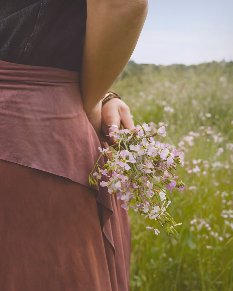Person in brown skirt holding small pink and white wildflowers behind their back in a grassy field.