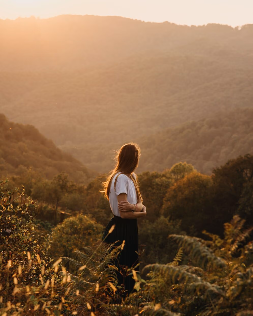 Woman in white top and black skirt standing in a sunlit forested area, looking at distant hills.