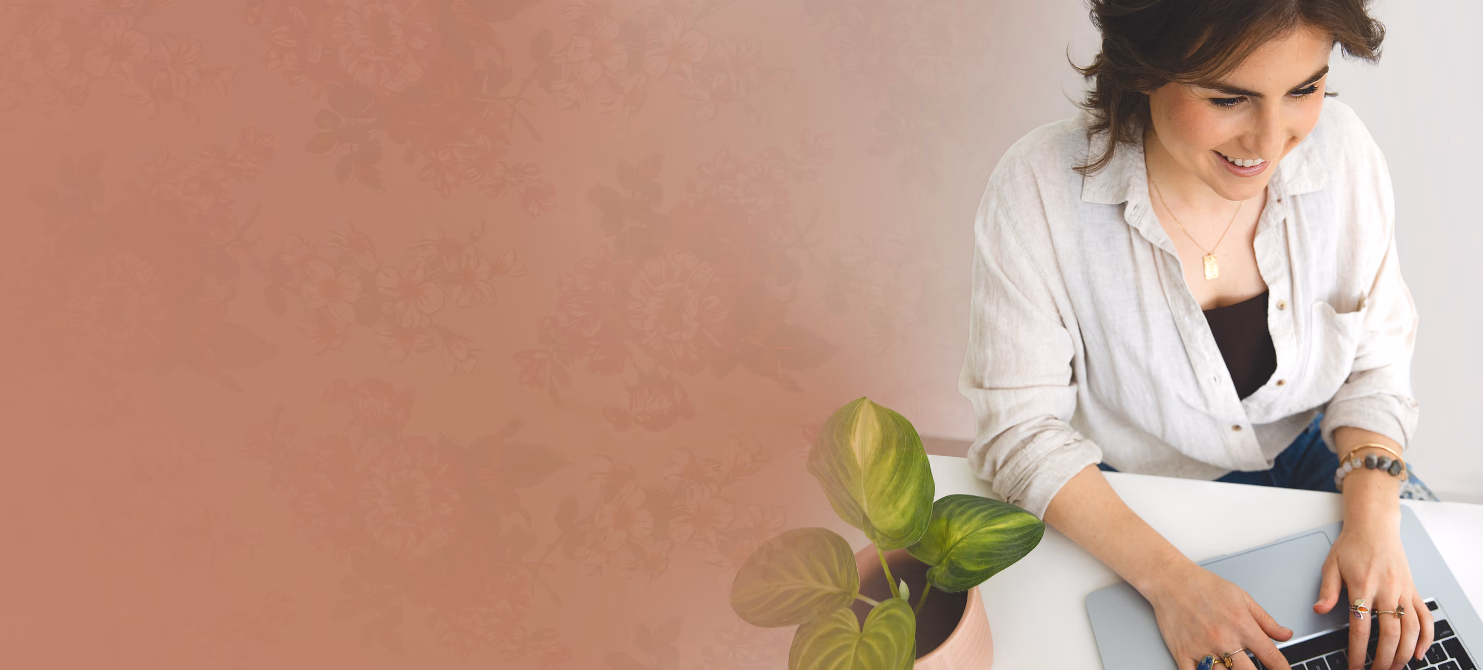 Leah Hoffman smiling and typing on a laptop at a white desk with a small green potted plant nearby.