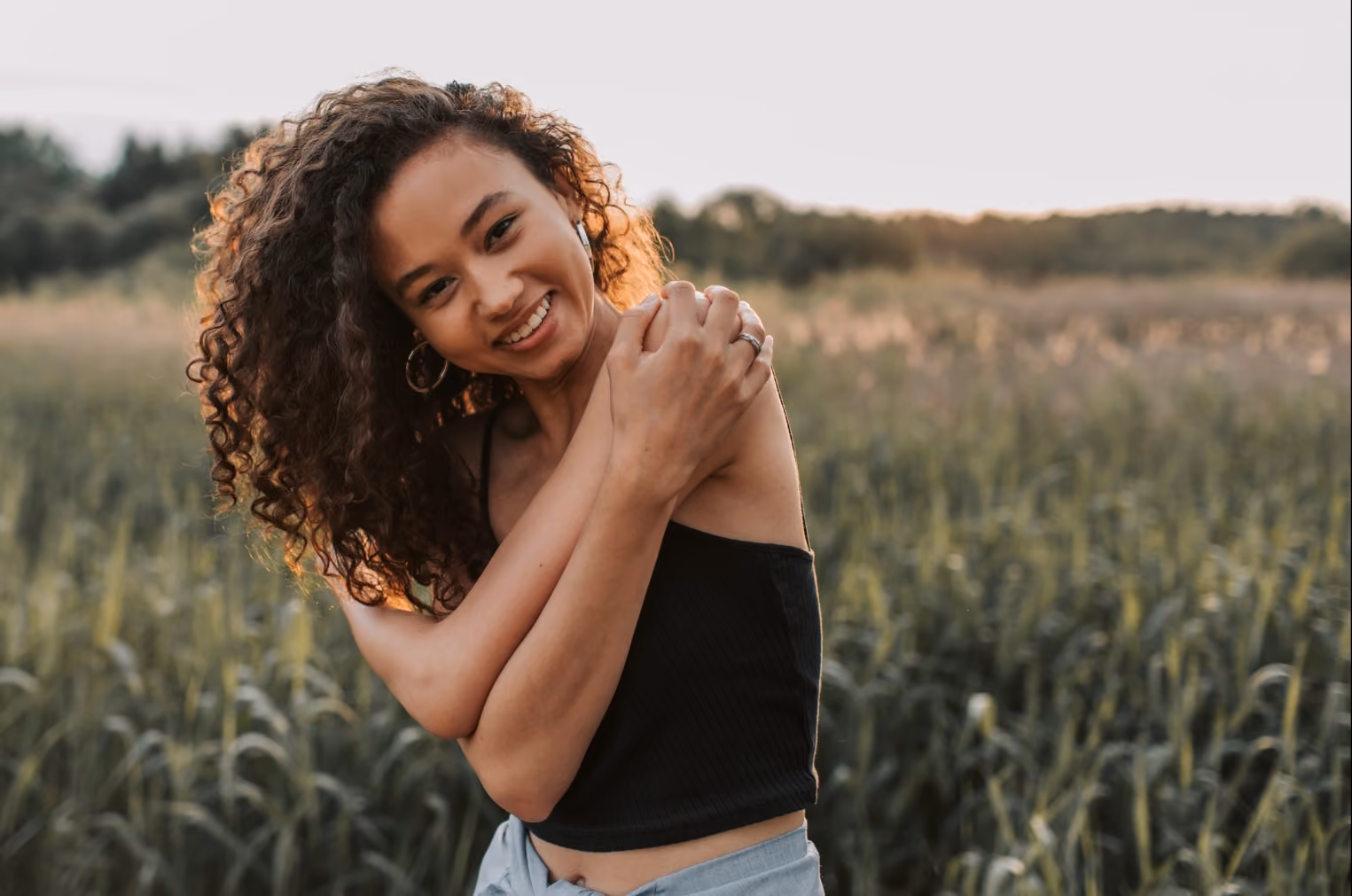 Smiling woman with curly hair in black top embraces herself in a green field at sunset.