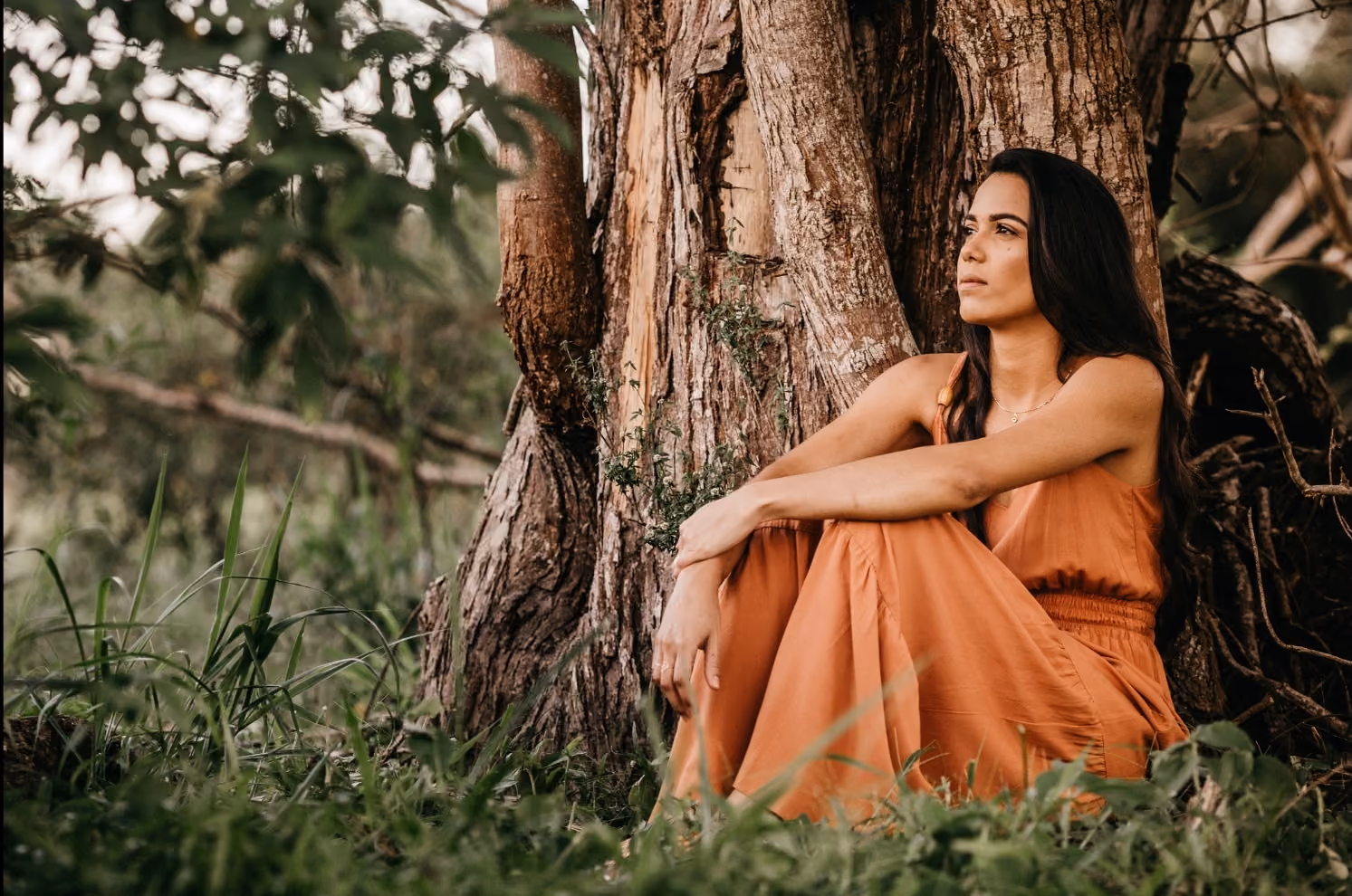 Woman in an orange dress sitting with arms around knees leaning against a large tree outdoors.