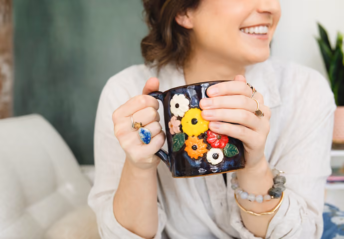 Leah Hoffman holding a dark floral-patterned mug while sitting on a white chair.