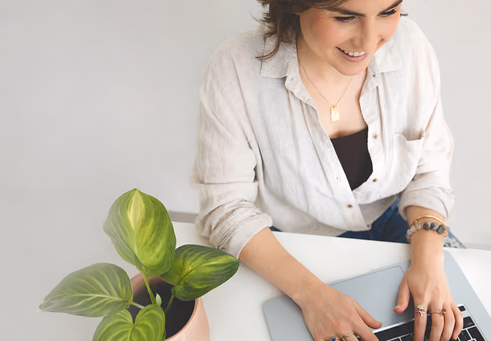 Leah Hoffman smiling and typing on a laptop at a white desk with a small green potted plant nearby.