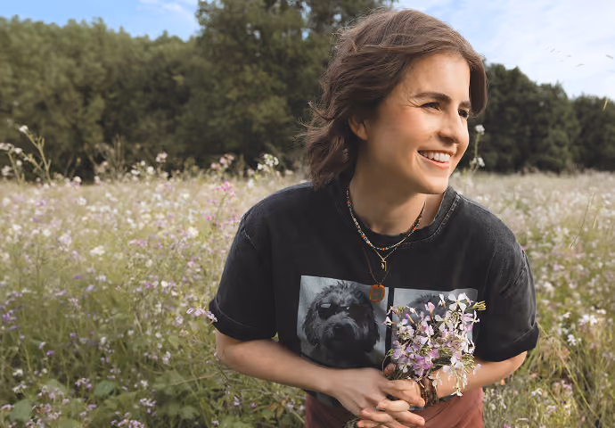 Leah Hoffman outdoors holding a small bouquet of wildflowers in a field with trees behind.