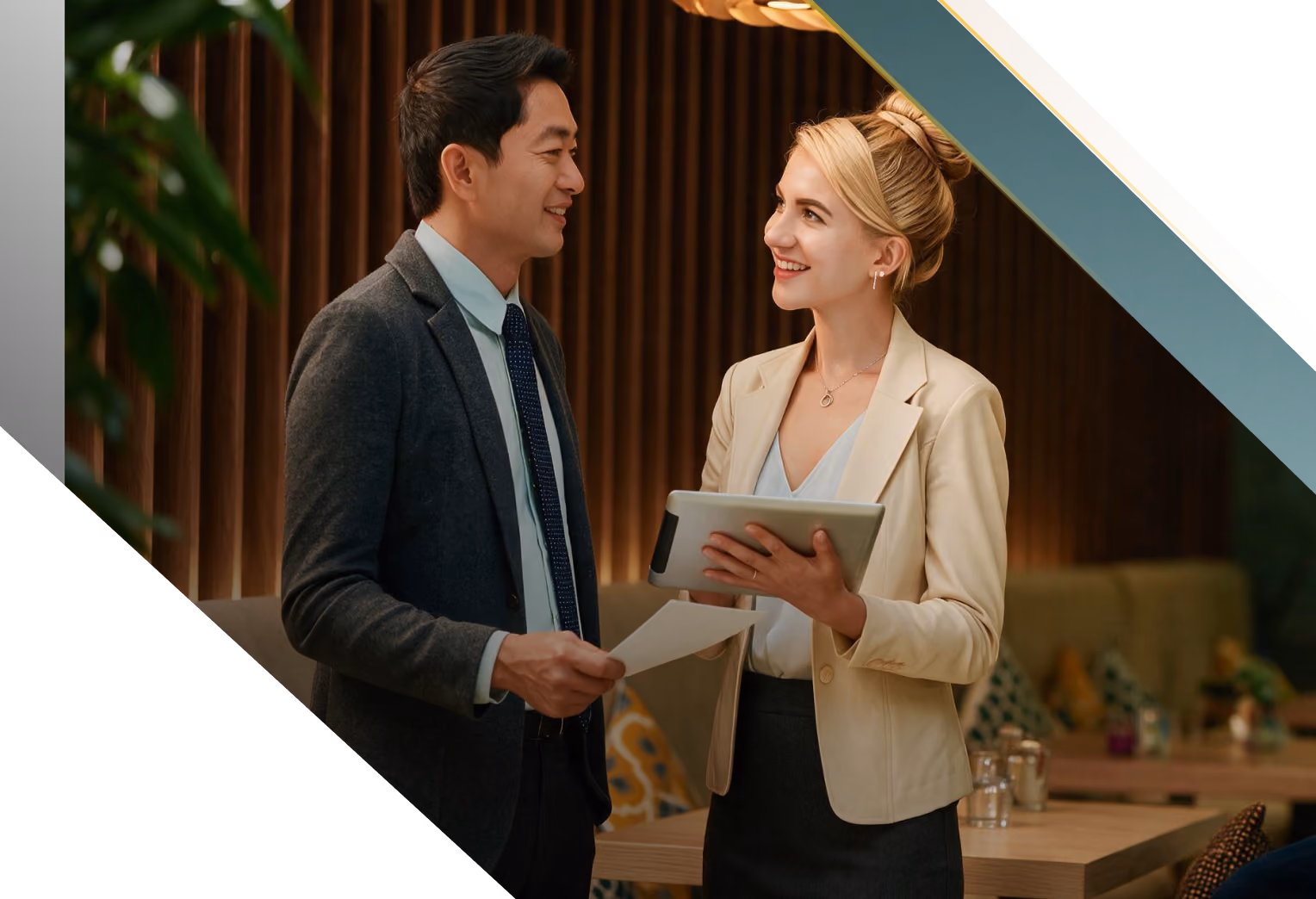 A man and a woman in business attire are smiling and having a conversation in a modern office or cafe setting, with the woman holding a tablet and the man holding a paper.