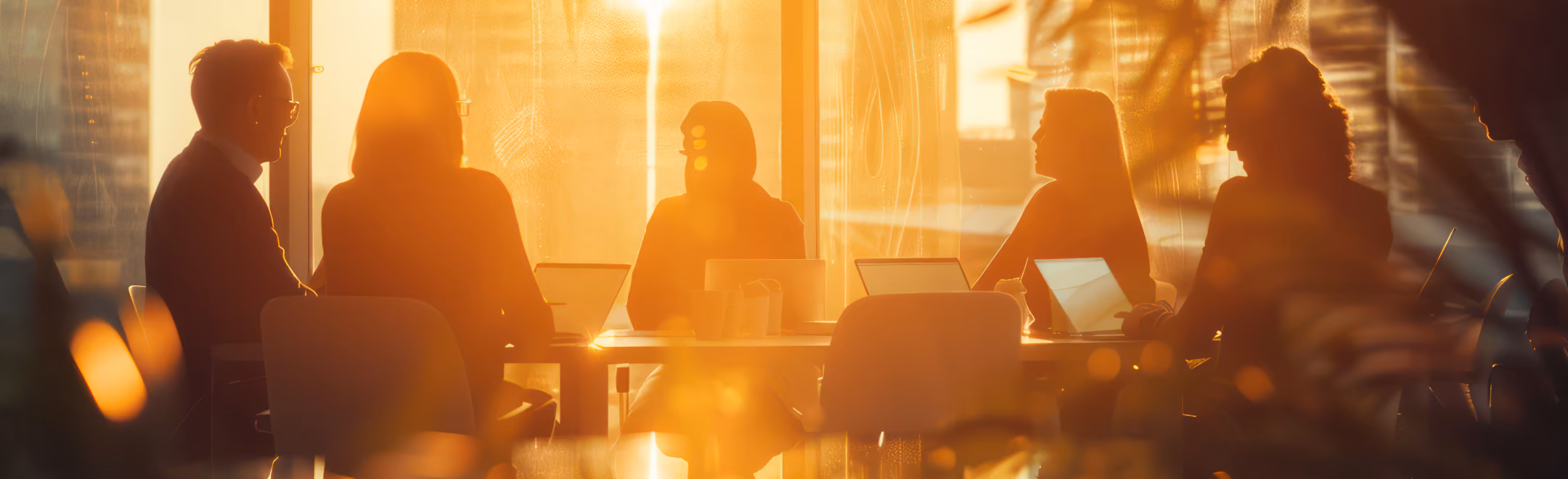 Silhouettes of six people sitting around a table engaged in a meeting or discussion with warm sunlight streaming through large windows behind them.
