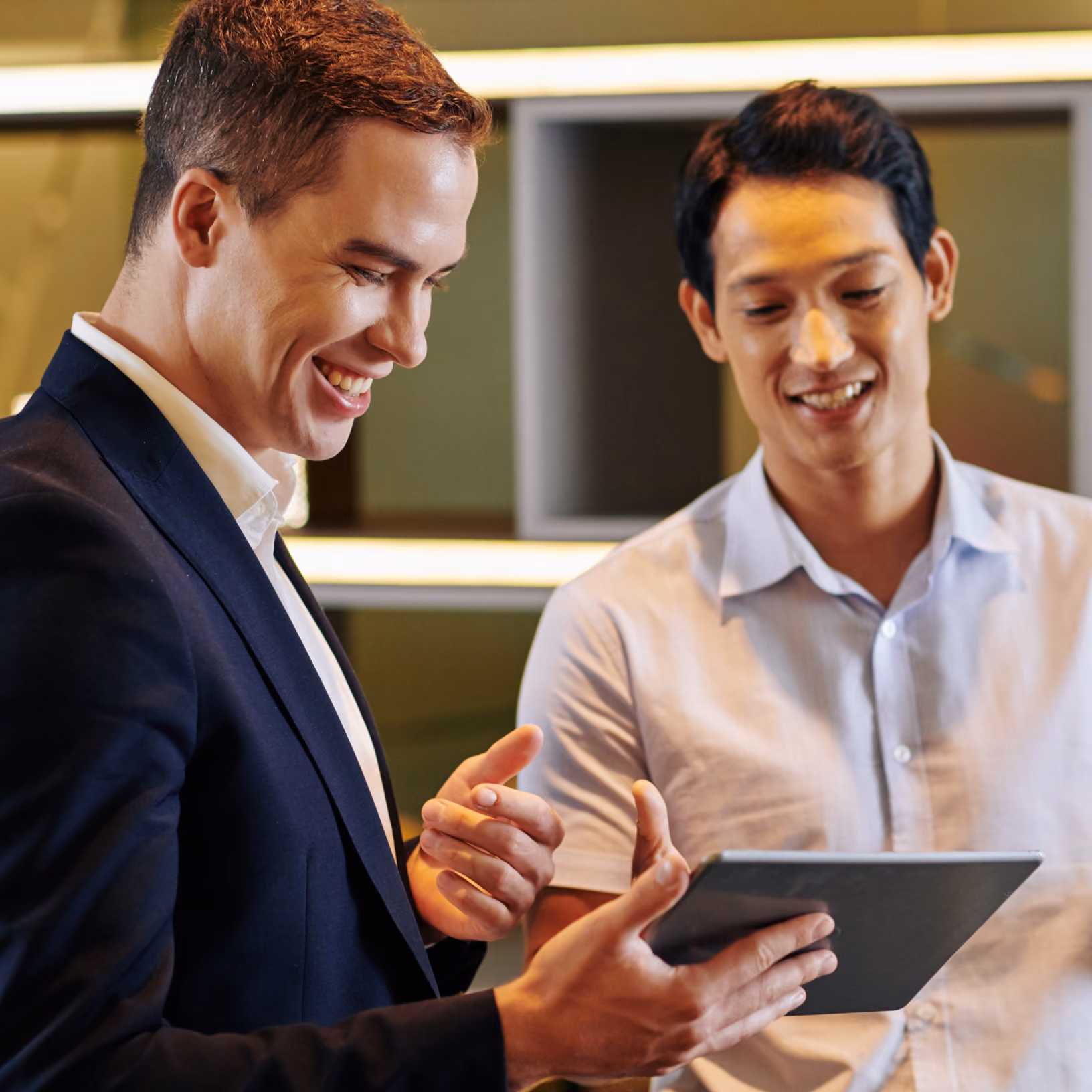 Two men smiling and looking at a tablet together, one in a dark blazer and the other in a light-colored shirt, in an indoor setting with modern decor.