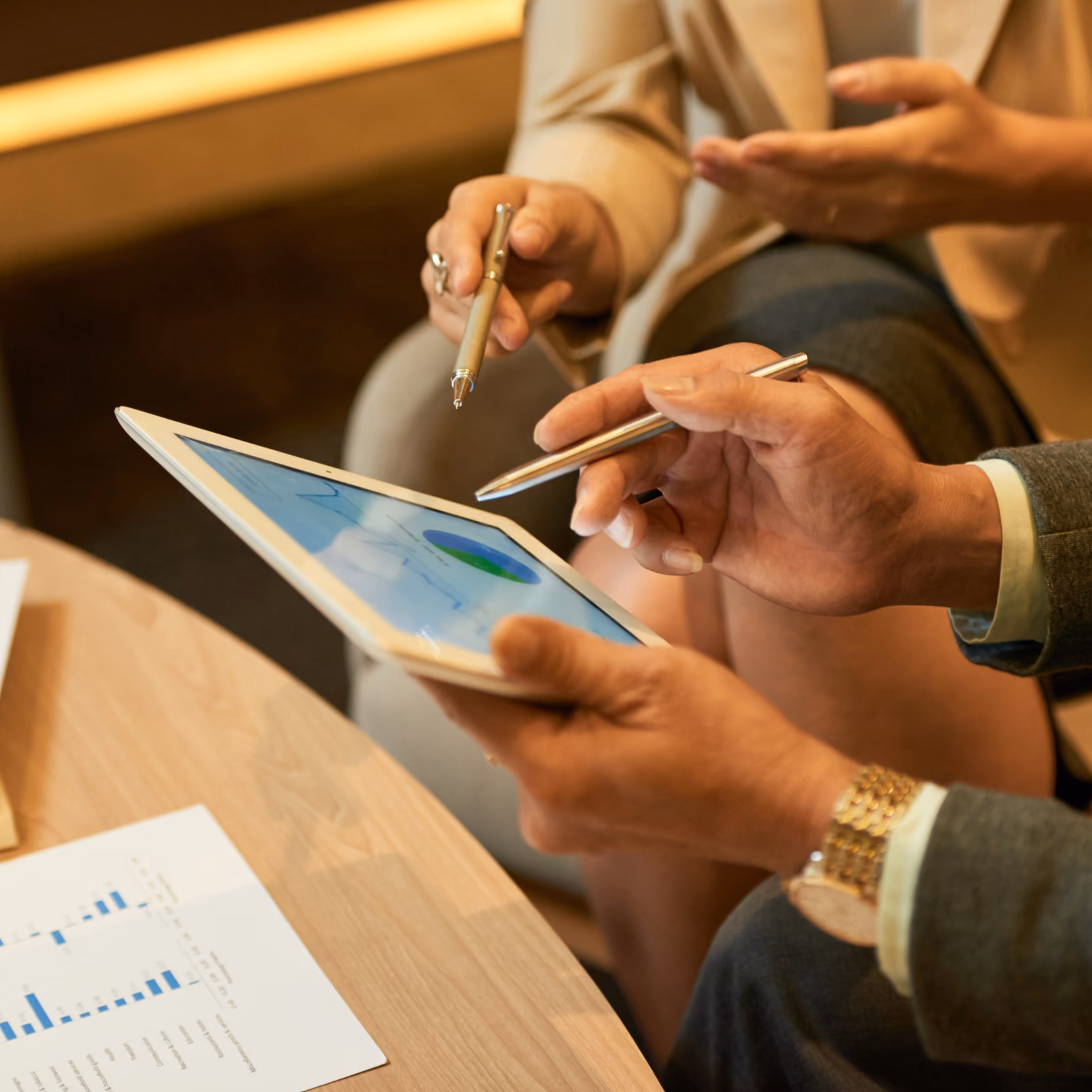 Two people using stylus pens to interact with a tablet displaying a colorful pie chart, sitting near a wooden table with a paper chart.