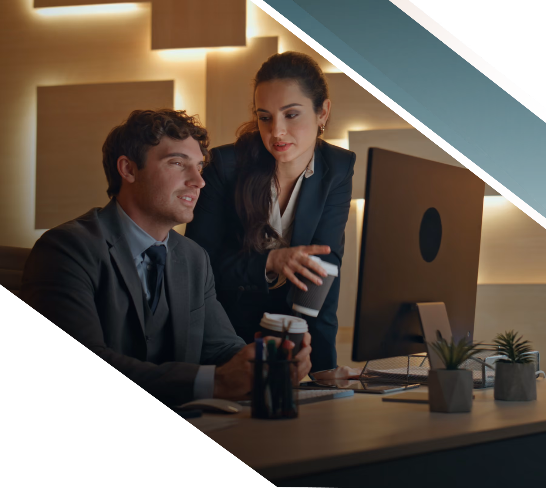 A man and a woman in business attire collaborate at a desk with a computer monitor, both holding takeaway coffee cups in a warmly lit office.