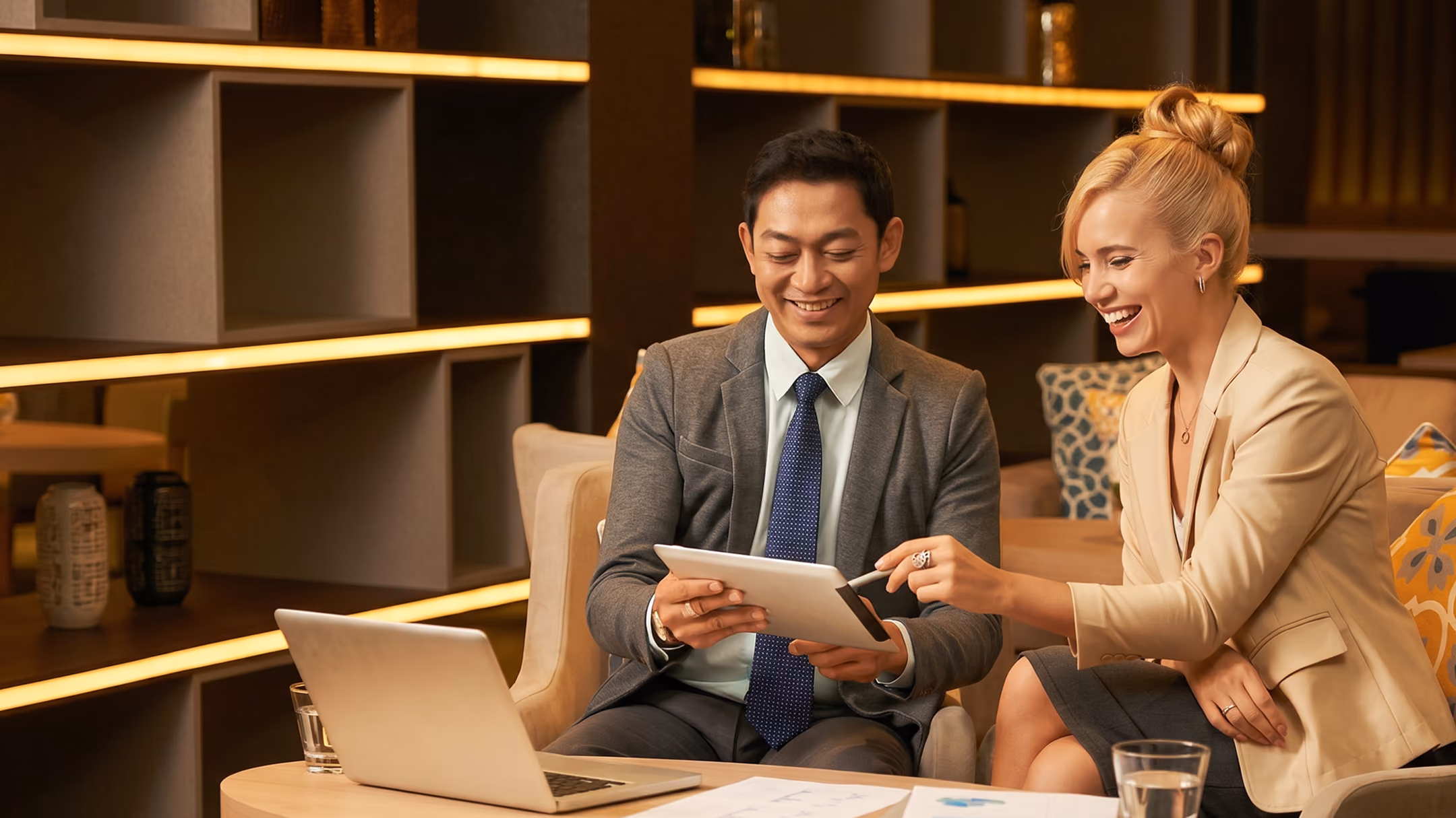 A man in a gray suit and blue tie and a woman in a beige blazer sit at a table with a laptop, smiling and looking at a tablet together in a warmly lit modern office lounge.