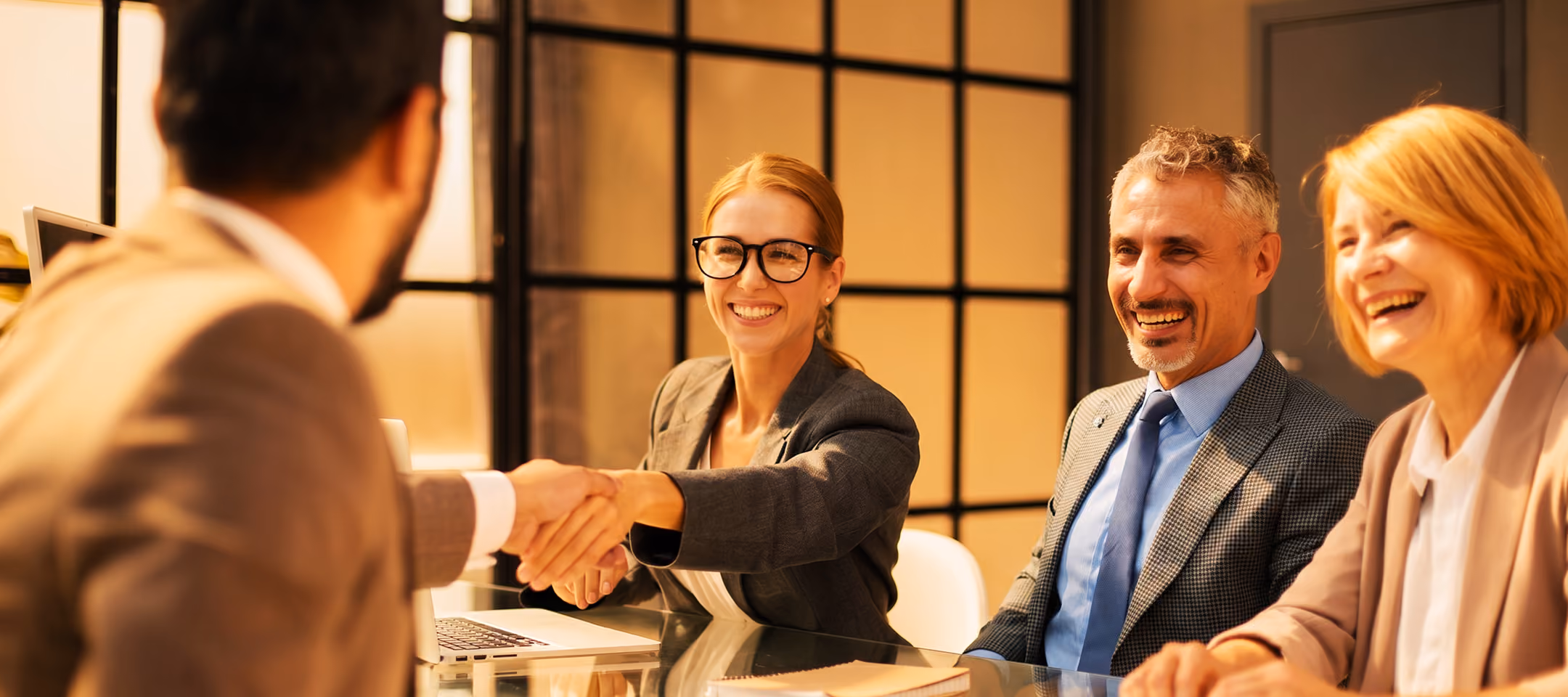 A businesswoman with glasses shakes hands with a man across a glass table as two colleagues smile and observe in a modern office setting.