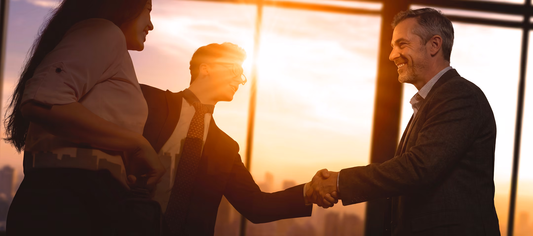 Two men in business suits shake hands and smile in an office with large windows during sunset, while a woman stands nearby.
